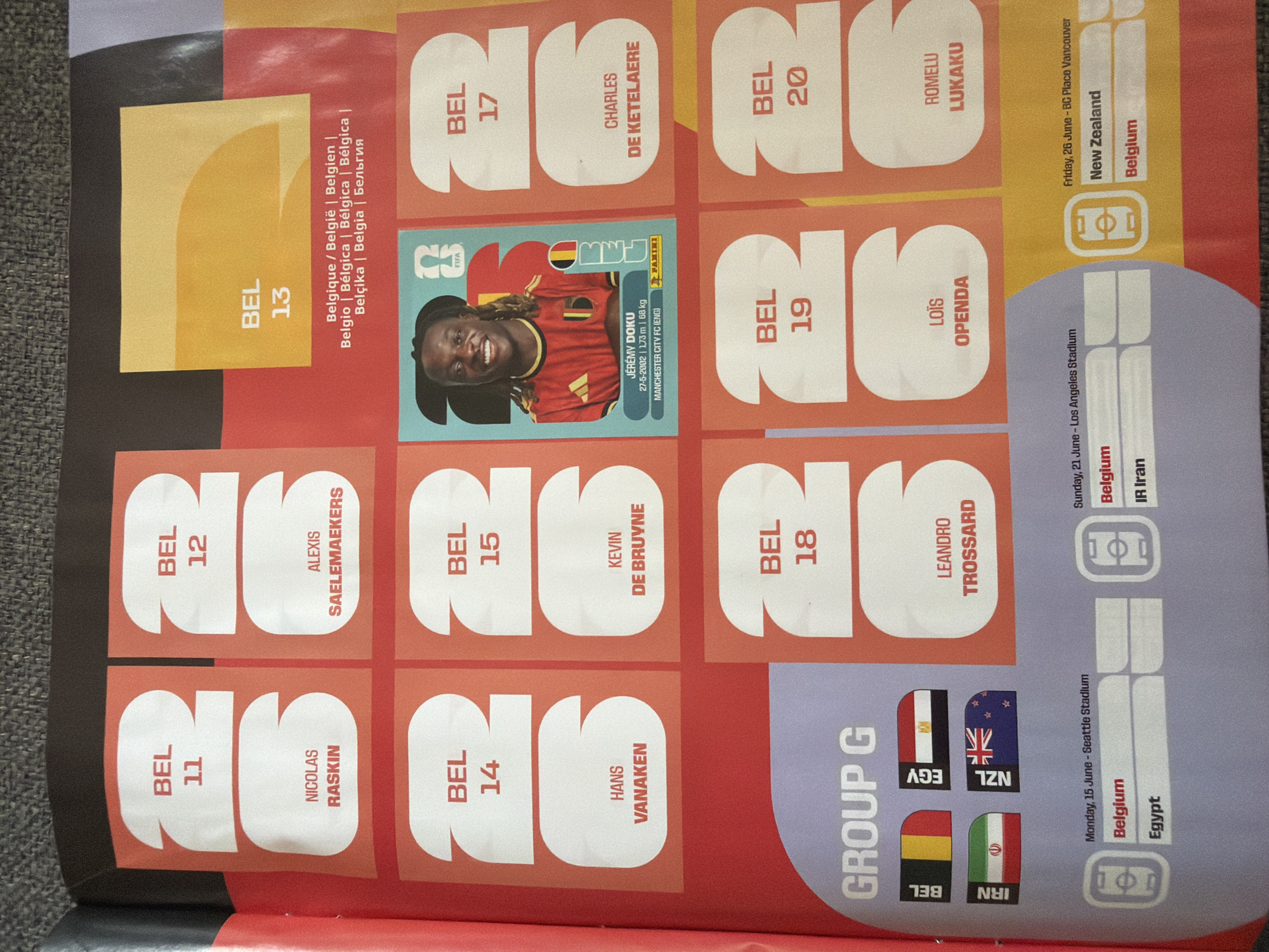 The image shows a page from a sticker album, likely related to a football (soccer) tournament. The page is divided into several sections, each featuring player stickers and team information.
In the top left section, there are two large stickers with the text "BEL 11" and "BEL 12" above spaces for player names. Below these are spaces for "NICOLAS RASKIN" and "ALEXIS SAELEMAEKERS".
To the right of this, a sticker for "BEL 13" is visible, with the player Jérémy Doku featured in a smaller sticker below it. The smaller sticker includes his name, birthdate, height, weight, club (Manchester City FC), and the FIFA and Panini logos.
Further to the right, there are additional large sticker outlines labeled "BEL 17", "BEL 18", "BEL 19", and "BEL 20". Below "BEL 17" is the name "CHARLES DE KETELAERE". Below "BEL 18" is "LEANDRO TROSSARD". Below "BEL 19" is "LOÏS OPENDA". Below "BEL 20" is "ROMELU LUKAKU".
In the bottom left quadrant, the text "GROUP G" is prominent. Below this are flags representing Belgium (BEL), Egypt (EGY), Iran (IRN), and New Zealand (NZL).
The bottom section of the page details match schedules. One entry lists "Monday, 15 June - Seattle Stadium" with fixtures between "Belgium" and "Egypt". Another entry shows "Sunday, 21 June - Los Angeles Stadium" with fixtures between "Belgium" and "IR Iran". A third entry, dated "Friday, 26 June - BC Place Vancouver", lists "New Zealand" and "Belgium".
The overall scene is a flat lay of a page from a sticker album on a textured surface, likely a carpet or fabric, which appears to be grey. The lighting suggests an indoor environment. There are no people or actions depicted, only static elements of the sticker album. The location of Zlatitrap, Bulgaria, is not depicted or implied by any elements within the image.
