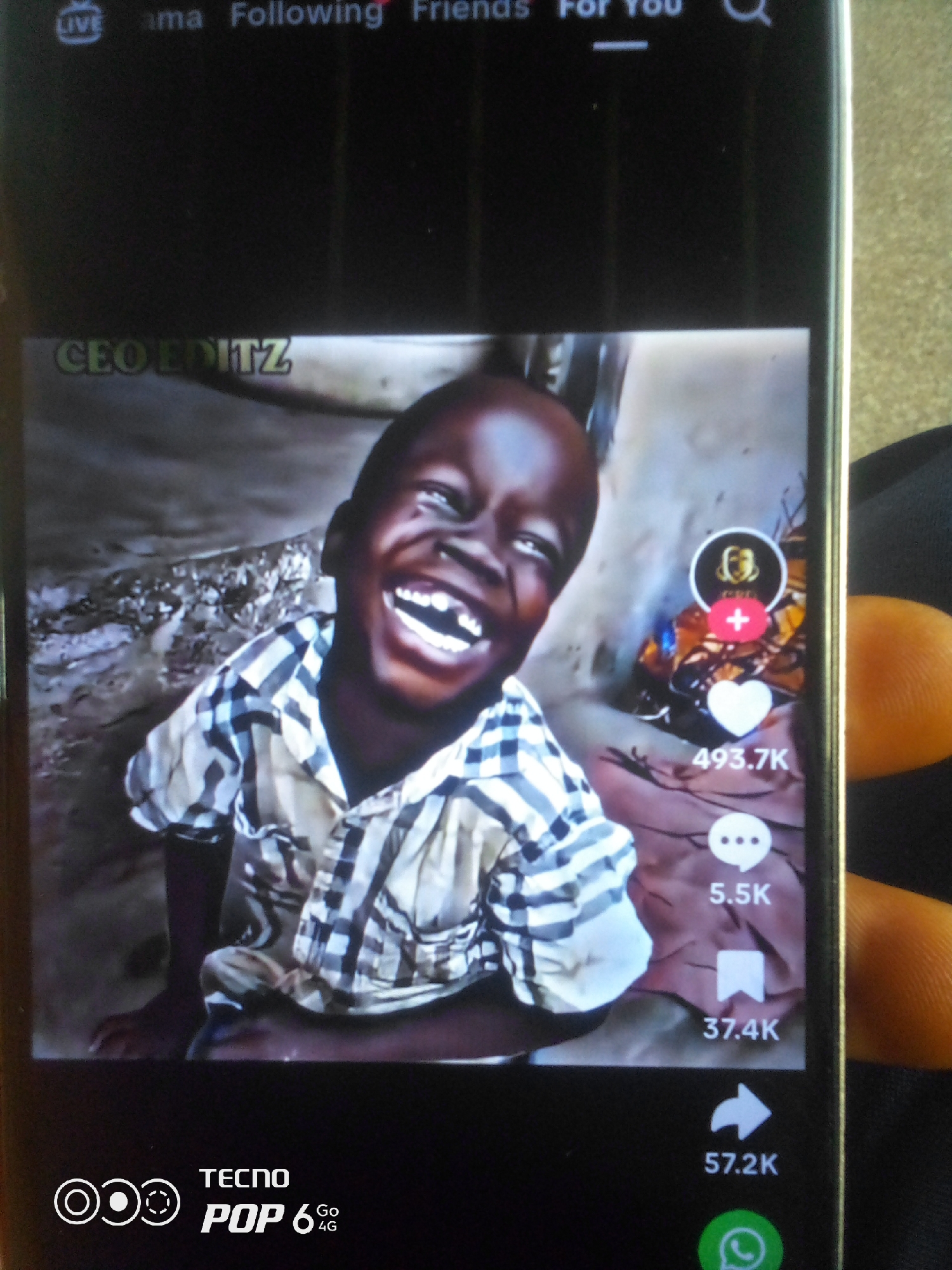 The image shows a screenshot of a video playing on a mobile phone. The video features a young Black boy with a wide smile and visible teeth. He is wearing a plaid shirt with black, white, and blue checks. The background of the video appears to be a rough, textured wall or ground. On the right side of the screen, there are icons for likes (heart symbol with "493.7K" below it), comments (speech bubble with "5.5K" below it), shares (flag symbol with "37.4K" below it), and a "share to WhatsApp" icon. The text "CEO EDITZ" is visible in the upper left corner of the video. The phone's brand, "TECNO POP 6 4G," is displayed at the bottom of the screen. The image is taken from an angle that includes a fingertip on the right side of the phone.