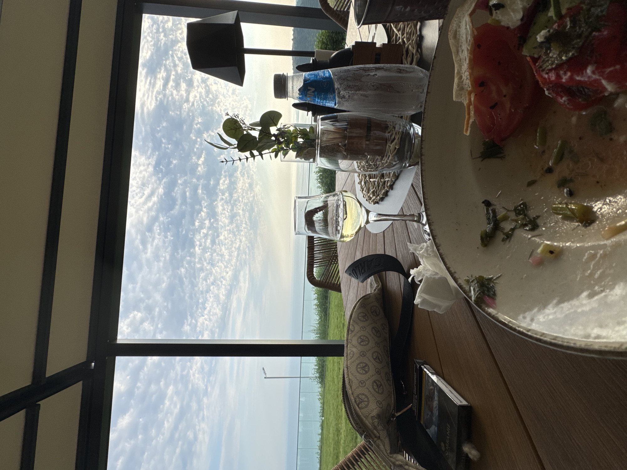 The image depicts a table setting at an outdoor restaurant overlooking a body of water and a grassy landscape. The perspective is from a low angle, looking up and to the right.
The table, made of wood, holds several objects. In the foreground, a partially eaten meal is on a plate. The meal appears to consist of red ingredients, possibly tomatoes, with some sauce and greenery. To the left of the plate, a wine glass containing a pale yellow liquid, likely white wine, is visible. Further left, there are two decorative glass containers: one holds a small plant with green leaves, and the other contains what appear to be wooden skewers or sticks. Behind these, a frosted water bottle with a blue label is placed on a woven placemat. A dark-colored purse or bag with a repeating pattern of interlocking circles and a strap is also on the table, positioned in the lower left quadrant of the image. A crumpled white napkin is near the plate.
Behind the table, a large window or glass partition offers a view of the outdoors. The sky is filled with dramatic, wispy white clouds against a bright blue backdrop, suggesting it is daytime, likely mid-morning or afternoon. Beyond the immediate greenery, a calm body of water is visible, with a distant shoreline or landmass. The overall atmosphere seems serene and possibly relaxed, with the meal and drinks suggesting a dining experience. The outdoor setting with the view hints at a pleasant day. There is a black lampshade visible in the upper left part of the image. A light pole is visible in the distance.
No people are visible in the image. The activity taking place is a meal being consumed or having recently been consumed, at a scenic location.
A portion of text is visible on a book or magazine placed on the table near the purse. The visible text reads "SZAKANIE".