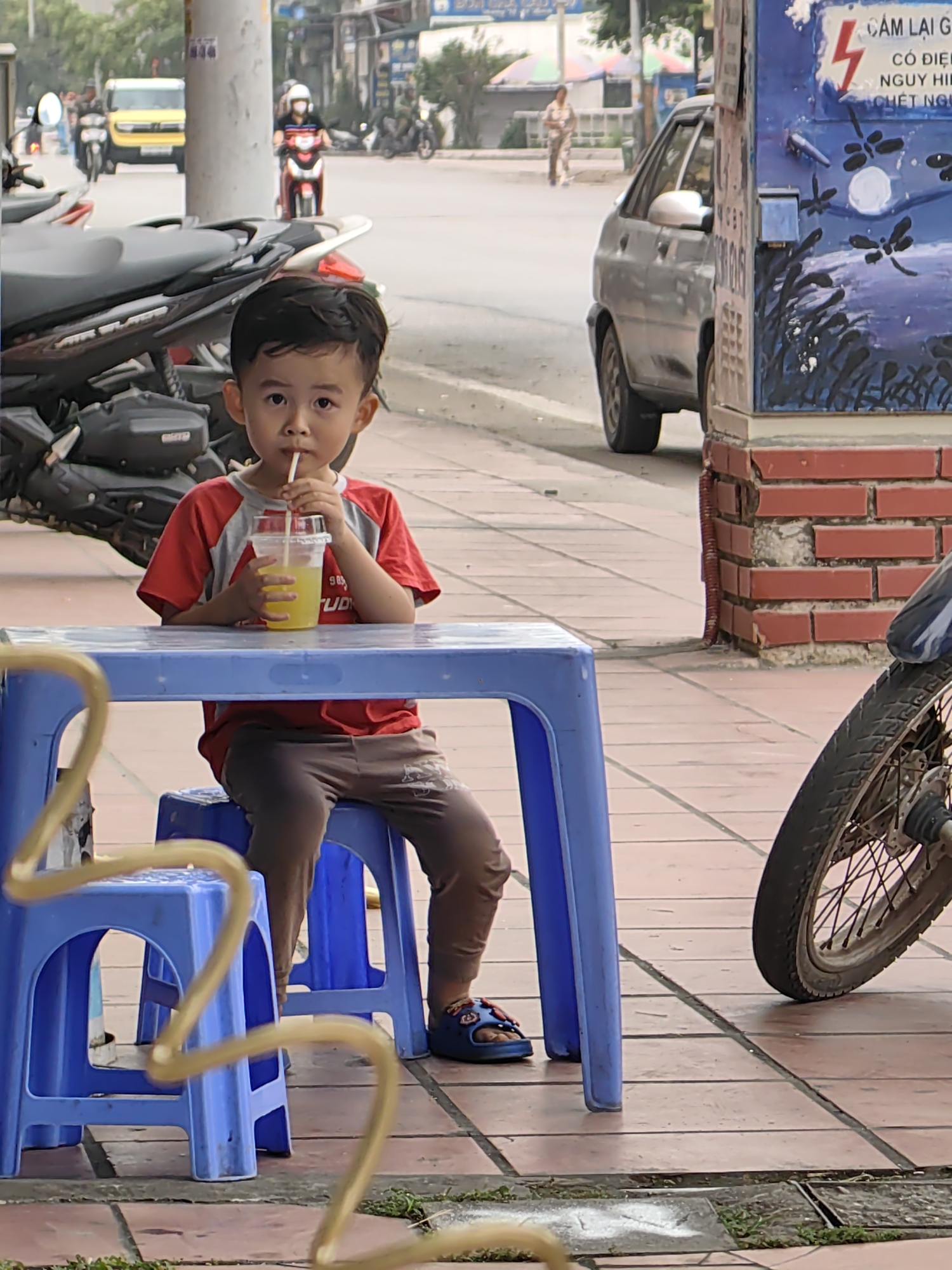 A young boy sits at a blue plastic table on a tiled sidewalk, drinking from a clear plastic cup with a straw. He is wearing a red and grey t-shirt, brown pants, and blue sandals. Behind him is a parked motorbike, and across the street, a yellow van and other motorbikes are visible. A parked silver car and a utility box painted with dragonflies stand to the right of the boy. In the background, a person walks along the street. A sign, partially obscured, is visible on the utility box. The scene appears to be located in Cao Xanh Ward, Vietnam, as indicated by the Vietnamese text on the sign.