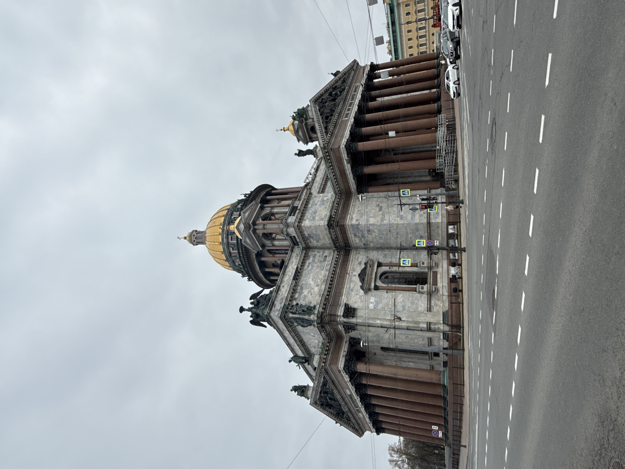 The image displays the magnificent Saint Isaac's Cathedral in Saint Petersburg, Russia, under an overcast sky. The grand Neoclassical edifice, positioned on the left side of the frame, features a prominent golden dome, topped with a smaller cupola and cross. The building's facade is adorned with numerous large, reddish-brown granite columns and intricately carved pediments embellished with sculptures and figures. Additional smaller gilded domes and sculptural elements are visible along the roofline.
To the right, a wide asphalt road stretches across the frame, marked by white dashed lines indicating multiple lanes. Several cars, including white and dark-colored vehicles, are parked along the far curb. In the background, beyond the road and parked cars, stands a light-colored building with green architectural details. Utility poles and overhead wires crisscross the upper part of the scene.
Near the base of the cathedral, beside the road, a traffic light pole displays a red light. Two pedestrian crossing signs (blue squares with white triangles and walking figures) are visible on this pole, along with a "No Stopping" sign (red circle with a diagonal line). A single, indistinct human figure can be seen near the traffic light, possibly a pedestrian. The sky is a uniform, somber gray, indicating an overcast day. Bare or sparsely-leafed trees in the lower right corner suggest cooler weather, possibly autumn or winter. The scene captures the cathedral as a dominant urban landmark.
