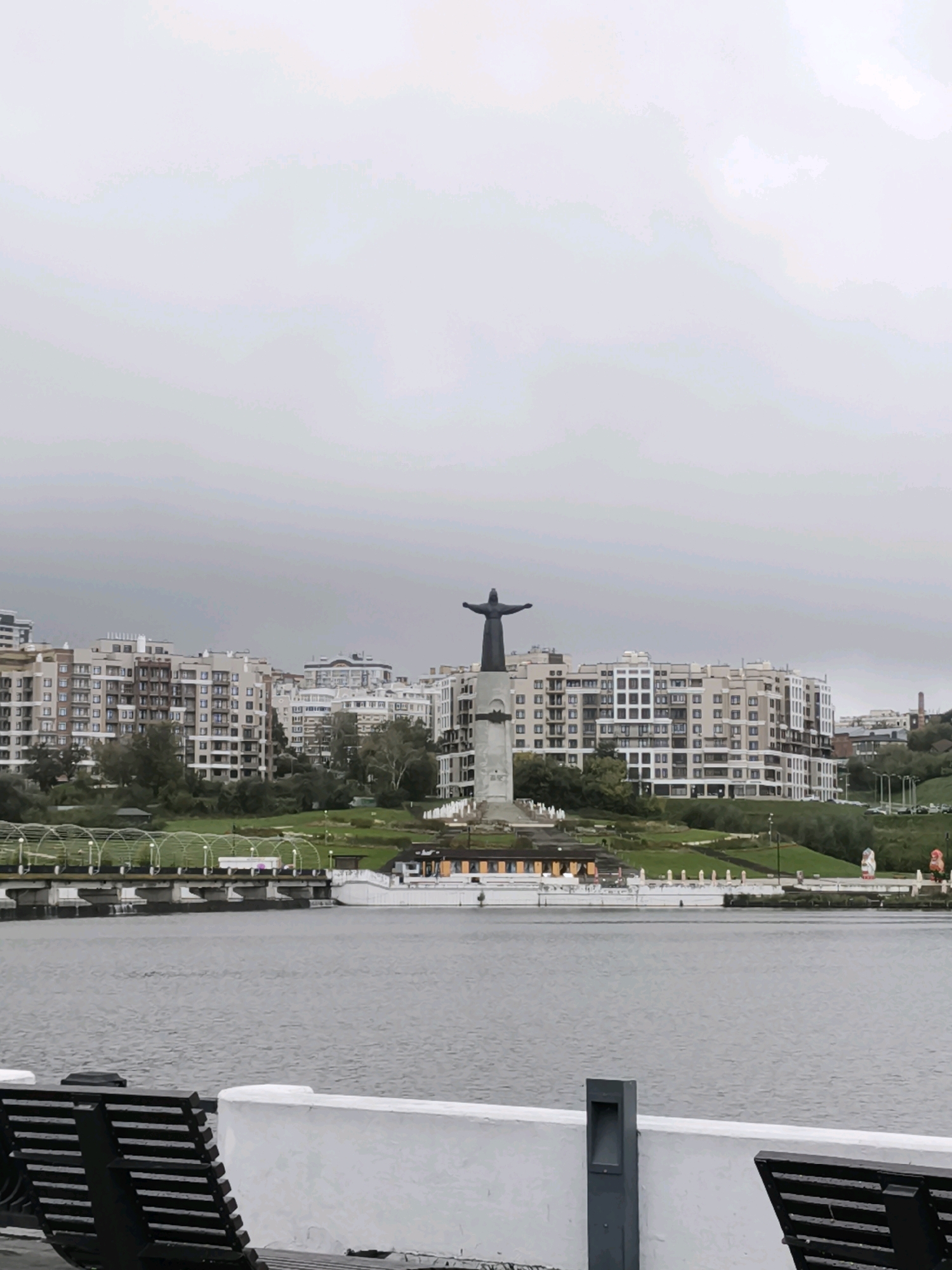 The image shows a wide shot of a waterfront in Cheboksary, Russia. In the foreground, there are several dark slatted benches and a white wall. Beyond the benches and wall, a large body of water fills the lower half of the frame. In the middle ground, a promenade or walkway runs along the water's edge, with a white structure that appears to be a pier or bridge. Behind the walkway, a green grassy slope leads up to a cluster of modern, multi-story apartment buildings. Dominating the center of the middle ground is a tall statue of a man with outstretched arms, standing on a cylindrical pedestal. The sky above is overcast and grey. The overall scene depicts an urban waterfront with a prominent monument and residential buildings.