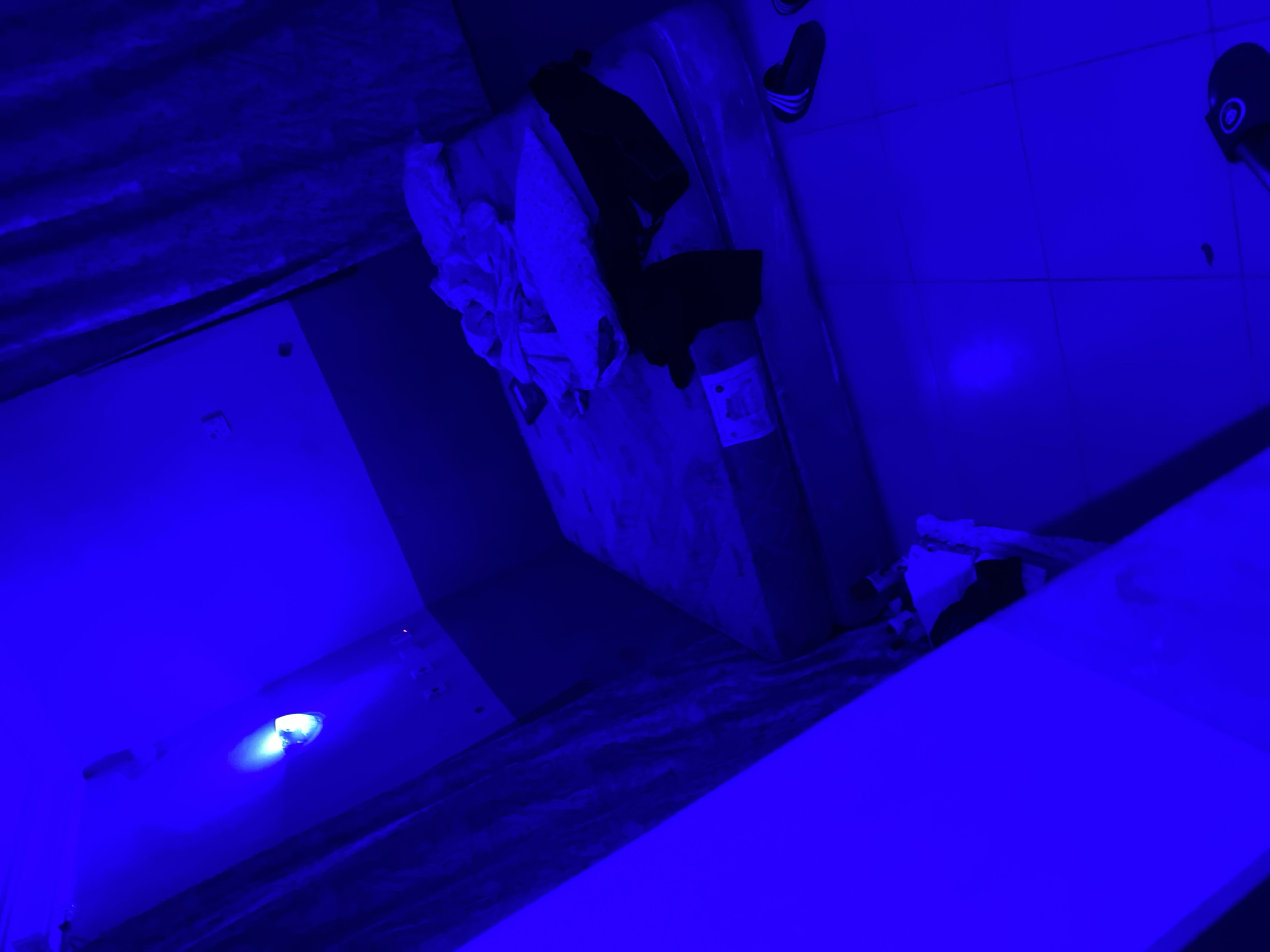 The image depicts a bedroom illuminated by a bright blue light, casting a neon glow over the entire scene. In the center of the room, a mattress lies on the floor, unmade, with several pillows and what appears to be dark clothing strewn across it. To the right of the mattress, a pair of black and white striped slides are visible on the floor. The walls are partially covered by dark curtains, and an electrical outlet is mounted on one wall. The overall impression is one of a dimly lit, personal space, possibly a bedroom, with a minimalist and somewhat unkempt presentation. The location in Howayi, Nigeria, is not discernible from the visual cues within the image.