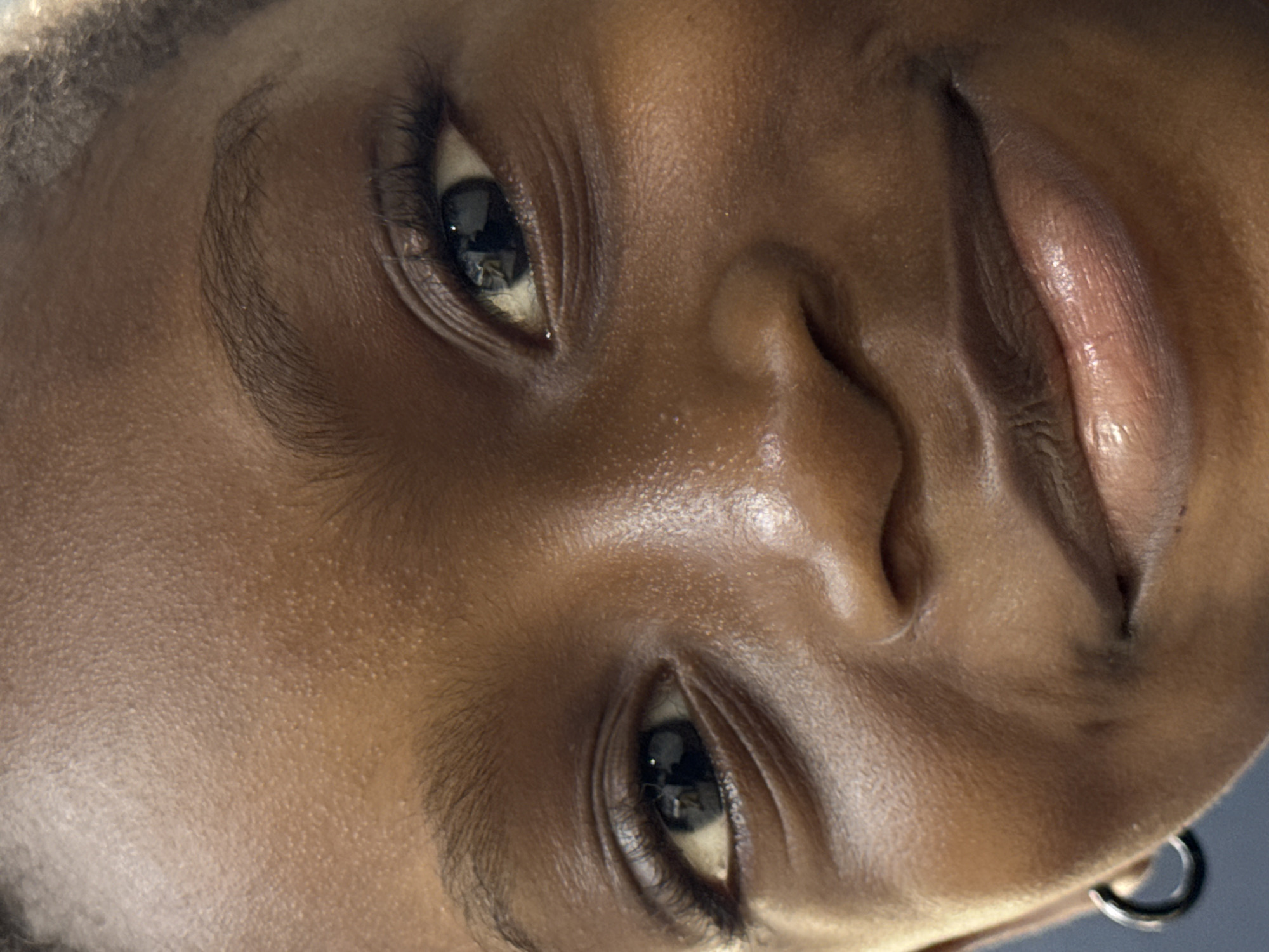 The image is a close-up, tilted portrait of a person with dark brown skin. The focus is primarily on the face, highlighting features such as the eyes, nose, and lips. The skin appears smooth and slightly glossy, with visible pores. The eyes are dark and reflect light. One earring is visible on the left side of the image. The background is blurred. No information is available to connect the subject to the city of Ado, Nigeria.