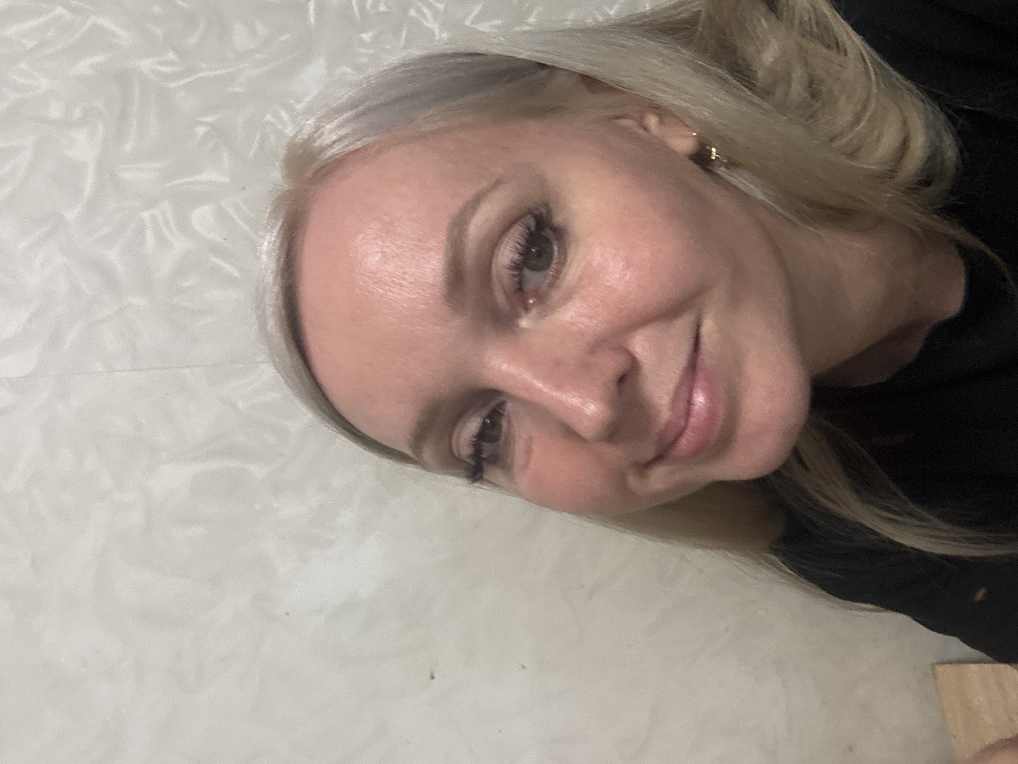 A blonde woman with light skin and green eyes is captured in a close-up, self-taken photograph. She is wearing makeup, including mascara and eyeshadow, and her hair is styled casually. The background consists of a textured, light-colored wall, which appears to be indoors. There are no discernible landmarks or specific features that would allow for identification of a location within the city of Komsomolsk-on-Amur, Russia. The image is a portrait focused solely on the woman's face and upper torso.