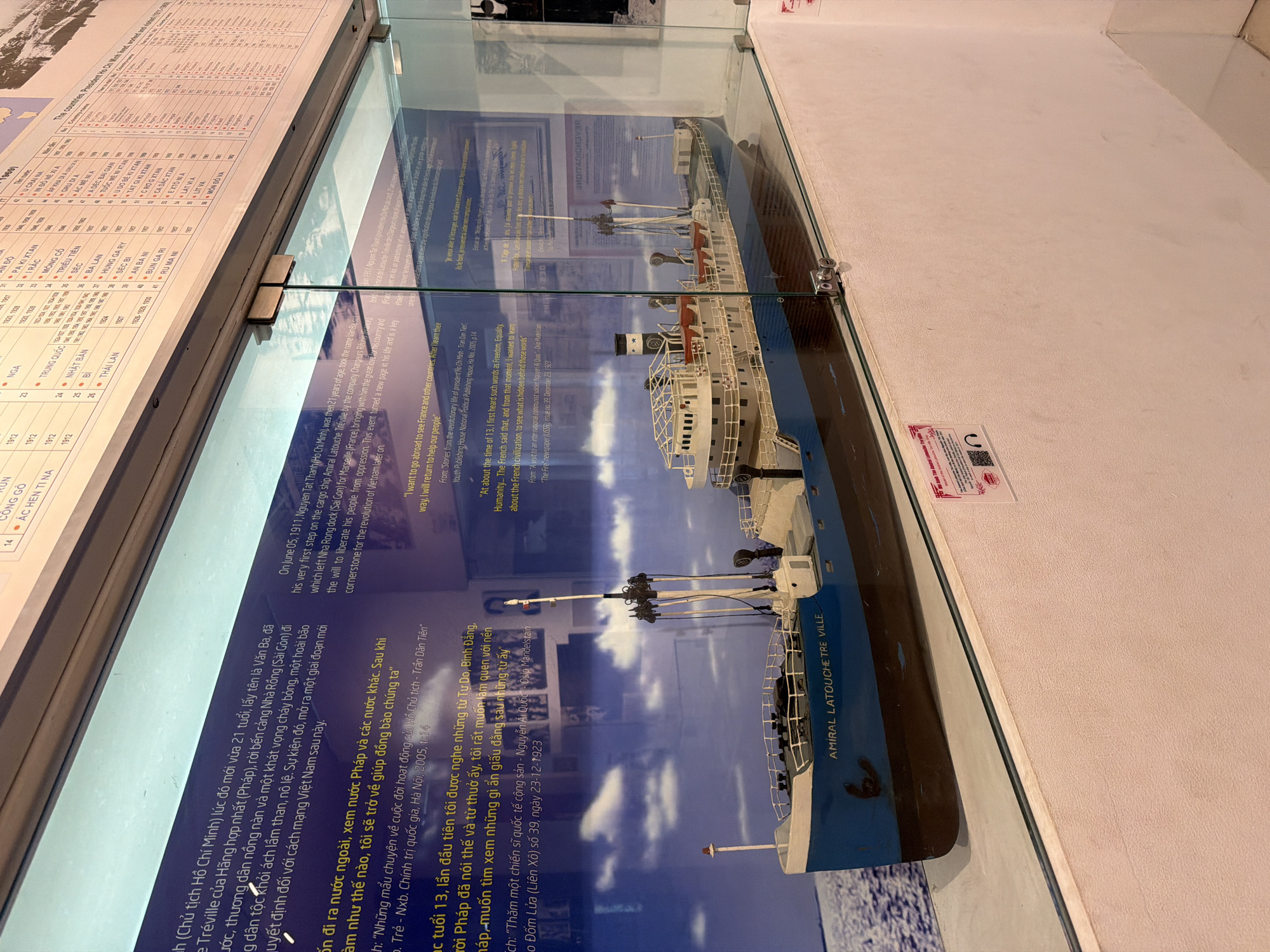 The image shows a model of the cargo ship "AMIRAL LATOUCHE TREVILLE" displayed in a glass case. The ship is painted blue and white, with several masts and decks. Behind the ship, a glass panel displays text in Vietnamese and French, with some English text at the bottom. The text appears to be historical information about the ship and its significance. To the left of the ship, there is a blue background with white clouds. To the upper right of the ship, there is a partially visible chart or list. The display is set against a textured wall, and the lighting suggests an indoor setting, possibly a museum or exhibition. The overall impression is a historical exhibit focused on maritime history. The location is stated as Thuận Hóa Ward, Vietnam.