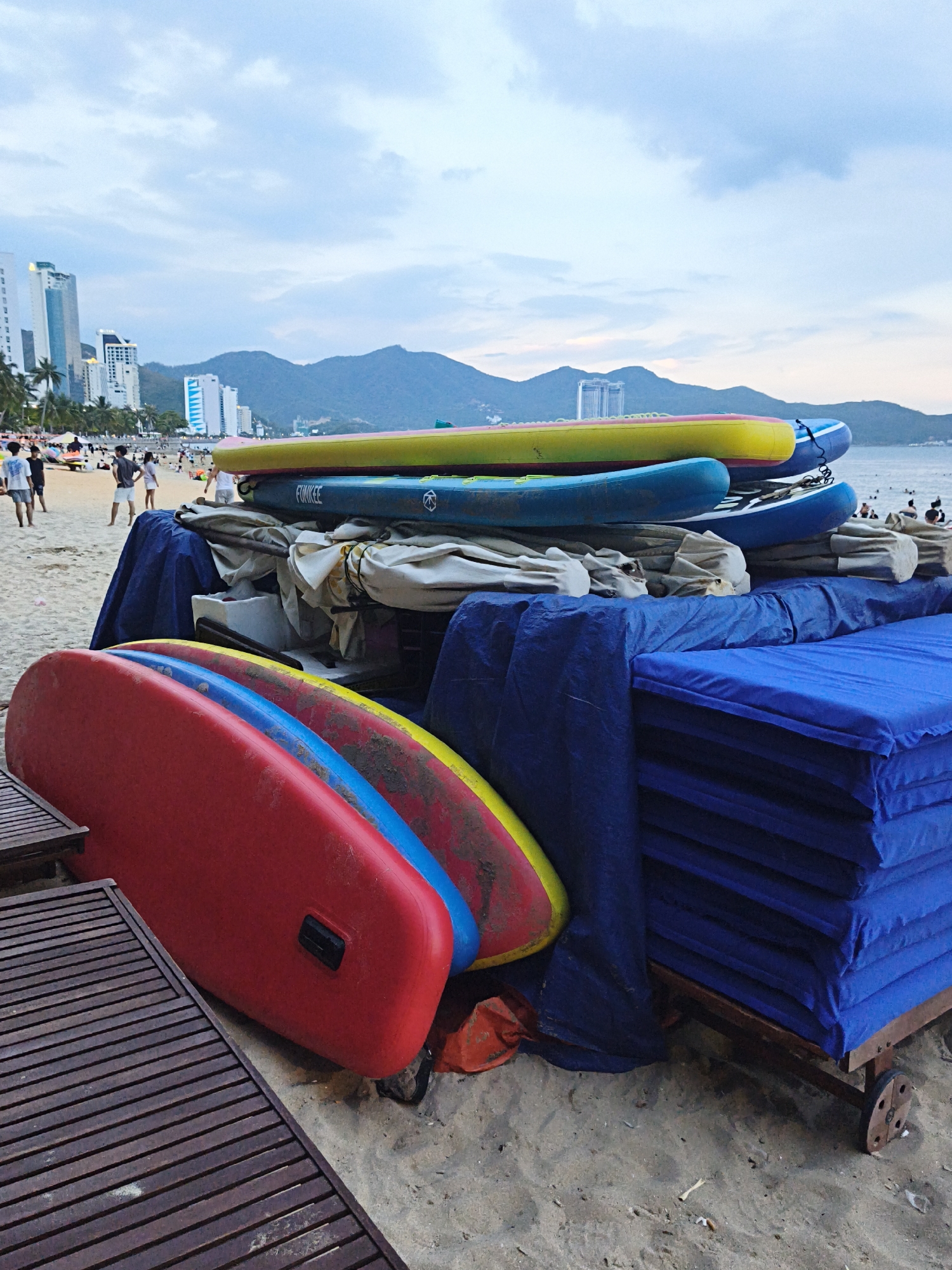 The image depicts a beachside scene in Nha Trang Ward, Vietnam, with a stack of colorful paddleboards and a cart laden with blue cushions. In the foreground, three paddleboards are visible: a red one on the bottom, followed by a blue and then a yellow one. They are resting on sand, with a wooden slatted table to the left. Behind the paddleboards, a large blue tarp covers a structure, and a cart with stacked blue cushions is partially visible to the right. The background shows a beach with scattered people, palm trees, and tall buildings, suggesting an urban coastal environment. In the distance, mountains are silhouetted against a cloudy sky.