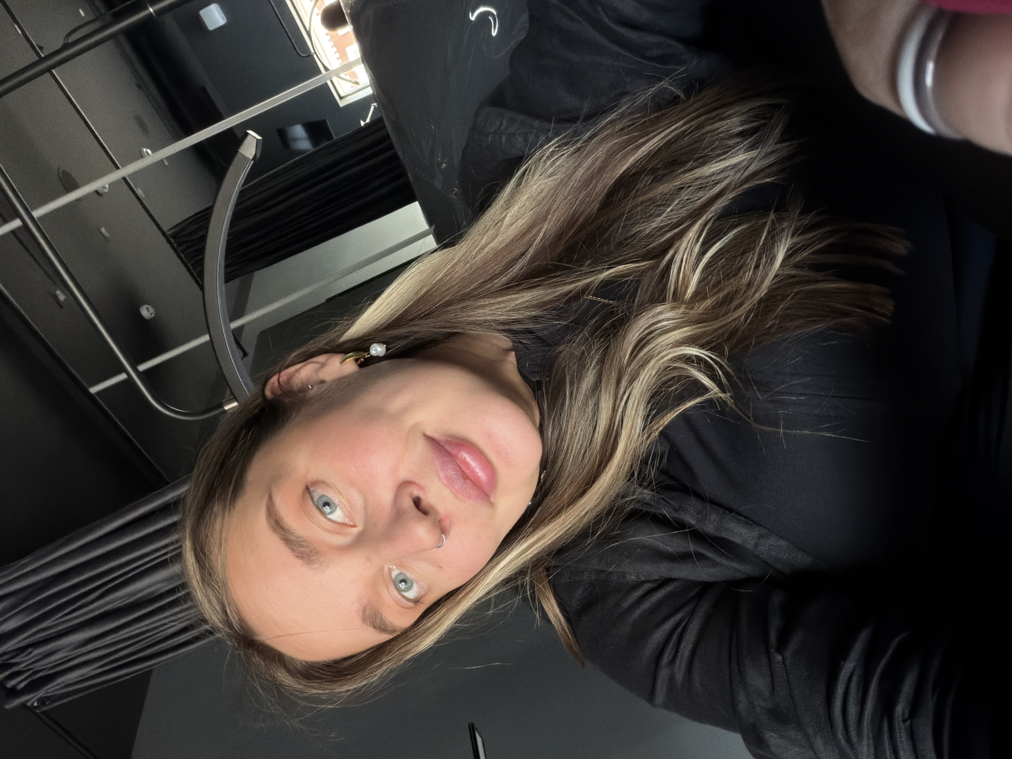 A woman with light brown and blonde hair, blue eyes, and a nose ring is posing for a selfie. She's wearing a black shirt. The background is predominantly dark, showing a ceiling with lighting fixtures and a curtain or fabric backdrop. The location cannot be determined from the image.