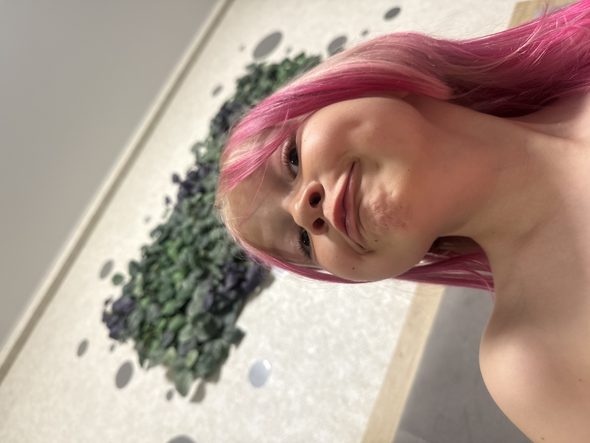 A close-up, slightly tilted photograph shows a person with pink hair. Their face is turned towards the camera with a slight smile. Behind the person, a wall is visible with a decorative plant arrangement and circular indentations. The location is Unknown, Unknown.