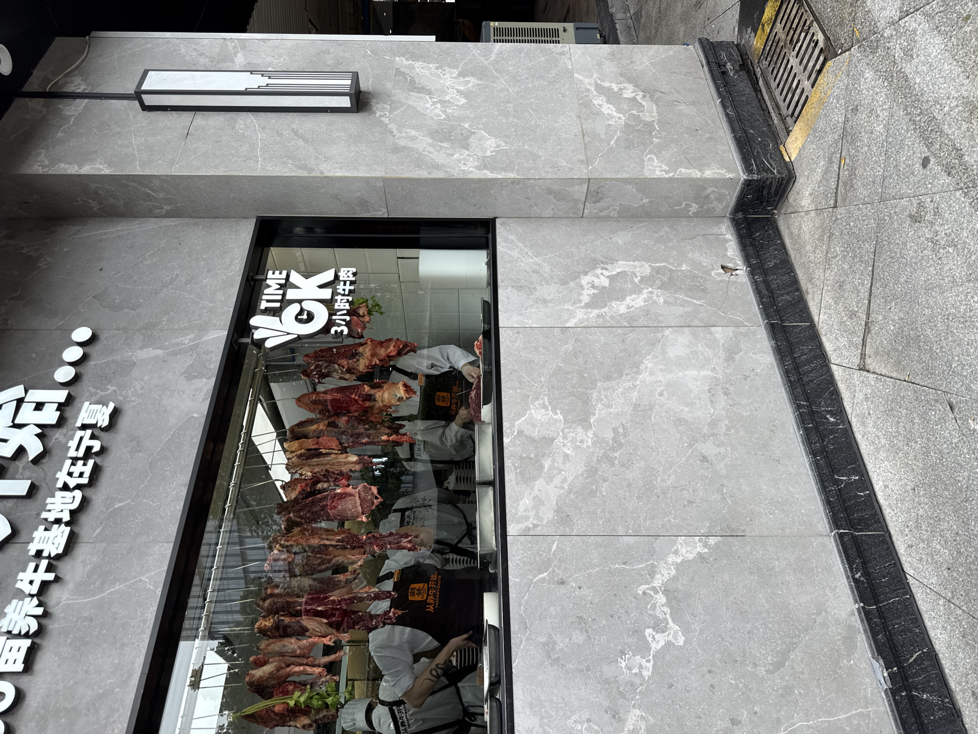 The image shows the exterior of a restaurant specializing in beef. The restaurant's name, "TIME OK" with Chinese characters below, is visible on the glass facade. Inside, raw beef carcasses are hanging, and several chefs in white uniforms and black aprons are preparing the meat. The restaurant is located on a street in Haizhu District, China, with a tiled facade and a drainage grate visible on the sidewalk. The overall impression is of a clean, modern establishment focused on high-quality meat.