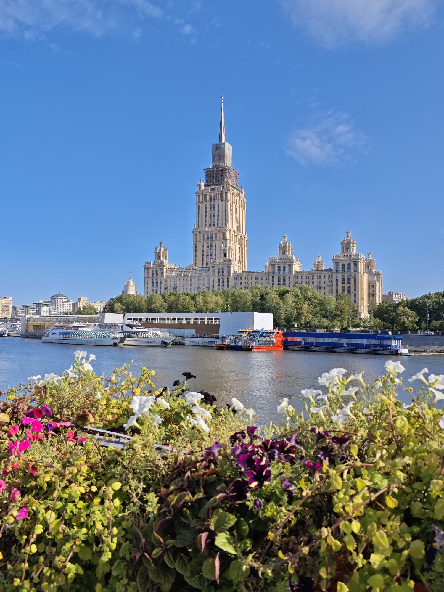 A bright, sunny day with a clear blue sky over Moscow, Russia. In the foreground, a vibrant flower bed filled with pink, purple, and white petunias and green foliage frames the scene. Beyond the flowers, the Moskva River flows gently, with several boats docked along the embankment. The most prominent structure in the background is the iconic Hotel Ukraina, also known as the Radisson Collection Hotel, a stunning Stalinist skyscraper with a distinctive spire. Other buildings and trees are visible along the river, creating a picturesque urban landscape. The water reflects the blue sky and buildings, adding to the serene atmosphere.