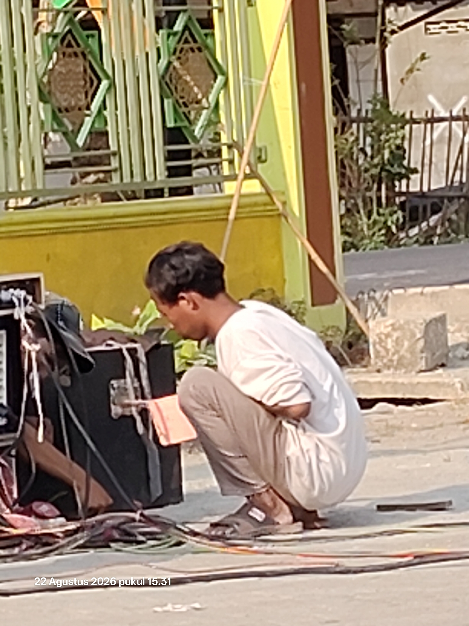 A man is crouching beside a large black speaker box in an outdoor setting, possibly a street or public area in Wedi, Indonesia, as indicated by the timestamp. He is wearing a white t-shirt and light-colored pants. His hands are near the speaker box, and he appears to be interacting with something on it, possibly adjusting cables or equipment. In front of the speaker box, there are numerous coiled cables and wires scattered on the ground, suggesting preparation or setup for an event. Behind the man and the speaker, there is a yellow building with green decorative elements, and further back, a fence with some vegetation. The ground is paved and appears to be a pathway or road. The overall scene suggests a temporary setup for audio equipment in a public space in the city of Wedi.