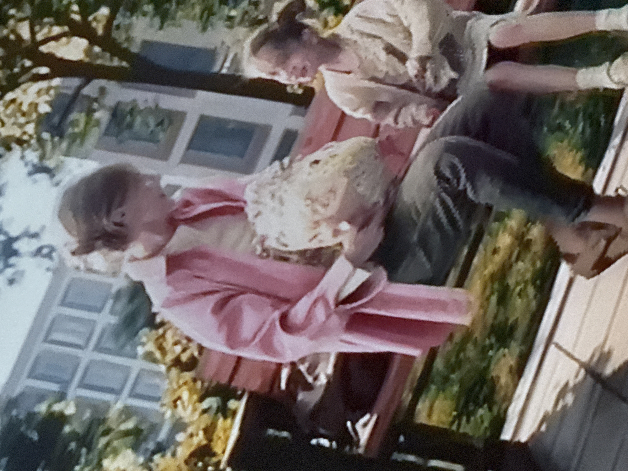 A photograph shows two people sitting on a park bench outdoors. One person, dressed in a pink coat, is holding a lacy white item in their lap. The other person is seated to their right. The setting appears to be a park or yard, with a building with multiple windows visible in the background. The photo's perspective is somewhat skewed. Without additional information, pinpointing the exact location within Mytishchi, Russia, is impossible.