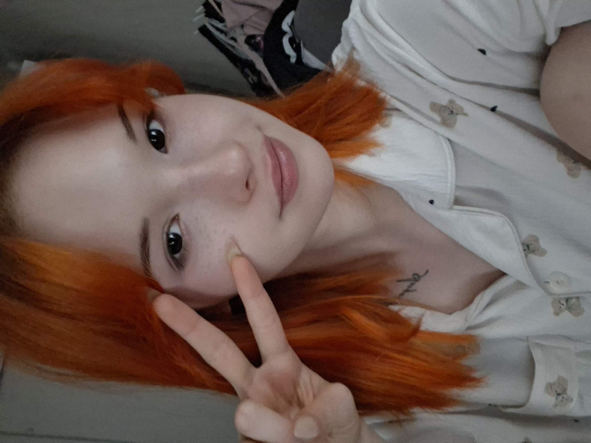 The image captures a close-up, slightly tilted selfie of a young person in an indoor setting, likely in Chaikovsky, Russia. The subject has striking, bright orange, shoulder-length hair and fair skin with visible freckles across their nose and cheeks. They possess dark, expressive eyes and full, naturally colored lips, with a subtle, gentle smile gracing their face.
The person is making a peace sign gesture with their left hand, holding it up near their face, slightly obscuring their cheek. They are wearing a light-colored, possibly white or cream-colored, collared shirt adorned with small, repeating teddy bear patterns and dark buttons. A small, stylized black tattoo is visible on their left chest/neck area. The tattoo appears to be a single word in cursive script, partially legible but difficult to decipher definitively; it resembles "love" or a similar short word.
The background is softly blurred and indistinct, suggesting an interior space like a room, with hints of darker elements behind them, possibly furniture or bedding. The lighting is soft and even, suggesting a daytime shot without harsh shadows. The overall mood conveyed by the subject's expression and gesture is calm and friendly.
