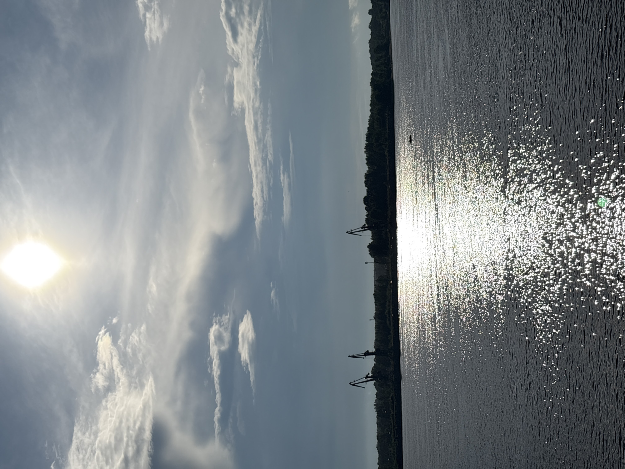 A wide shot captures the Kama River in Chaikovsky, Russia, under a bright, partly cloudy sky. The sun, a brilliant white disc, shines from the upper center, casting a shimmering reflection across the water's surface. The river takes up the lower half of the frame, its dark water dappled with countless points of light from the sun's glare. On the far bank, a dark line of trees forms the horizon, broken by the silhouettes of several tall industrial cranes, suggesting a port or industrial area. In the middle distance, a small boat with at least one occupant can be seen on the water. The sky above is a mix of pale blue and white clouds, some wispy and others more solid, with the sun piercing through. The overall impression is one of a serene, sunlit day by a major waterway, with hints of industrial activity in the background.