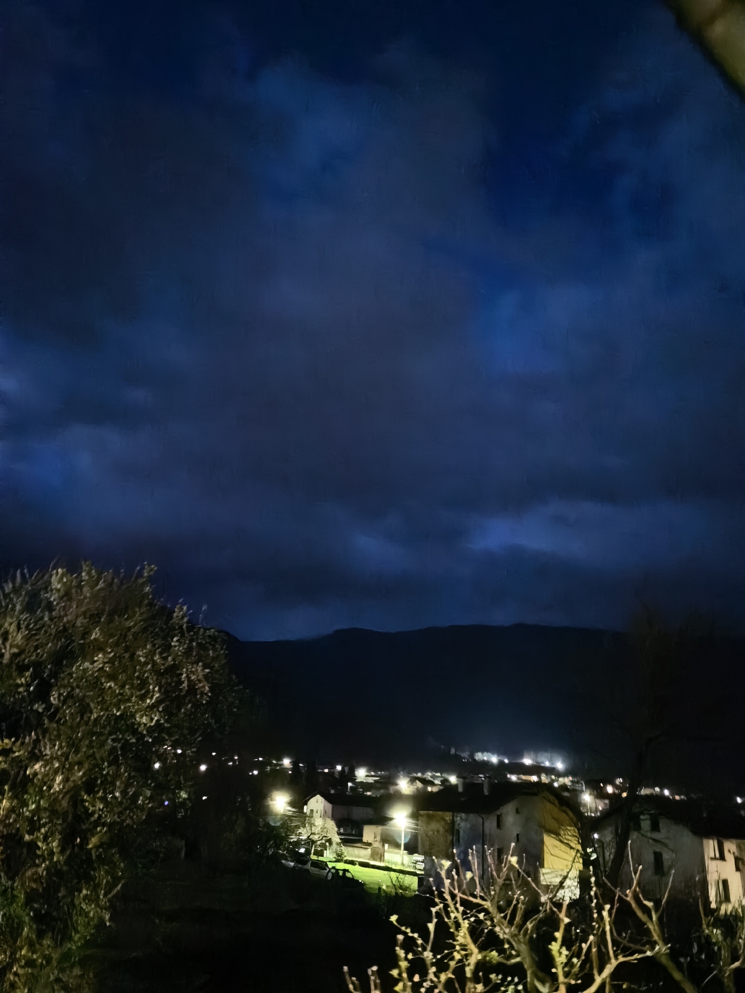 A nighttime outdoor shot depicts a landscape, with a town or village nestled in front of a dark mountain range. The sky is predominantly covered with dark blue, clouded sky. In the foreground, trees and vegetation are visible, framing the scene. The village below is illuminated by streetlights and lights emanating from buildings, casting a warm glow against the night sky. The buildings appear to be houses or low-rise structures. Cars can also be seen around the houses. The image appears to be taken from an elevated vantage point, possibly a hill or upper floor of a building. The location is Unknown, Unknown.
