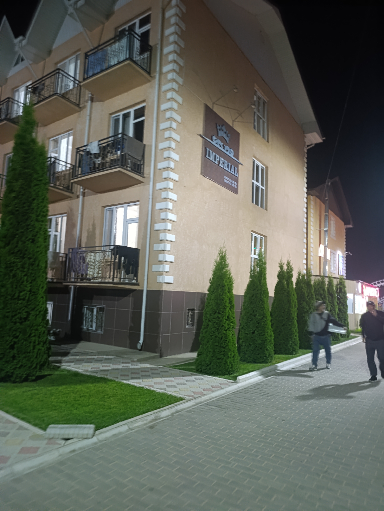 The image captures an exterior view of a multi-story building in Bosteri, Kyrgyzstan, at night. The building, possibly a hotel or residential complex, features a light brown facade, numerous balconies with black railings, and white decorative trim along its corners. A prominent sign reading "IMPERIAL" is visible on the upper facade, accompanied by a graphic of a crown. The building is flanked by a row of well-maintained green cypress trees and a manicured lawn. A paved walkway with a patterned brick surface stretches along the building's side. Two individuals are walking away from the camera on this walkway. The person in the foreground, on the left, is wearing a grey hooded jacket and blue jeans, carrying a white object. The person on the right is wearing a dark jacket and dark pants. The scene is illuminated by artificial light, casting long shadows from the trees and people onto the walkway. The background is dark, indicating nighttime.