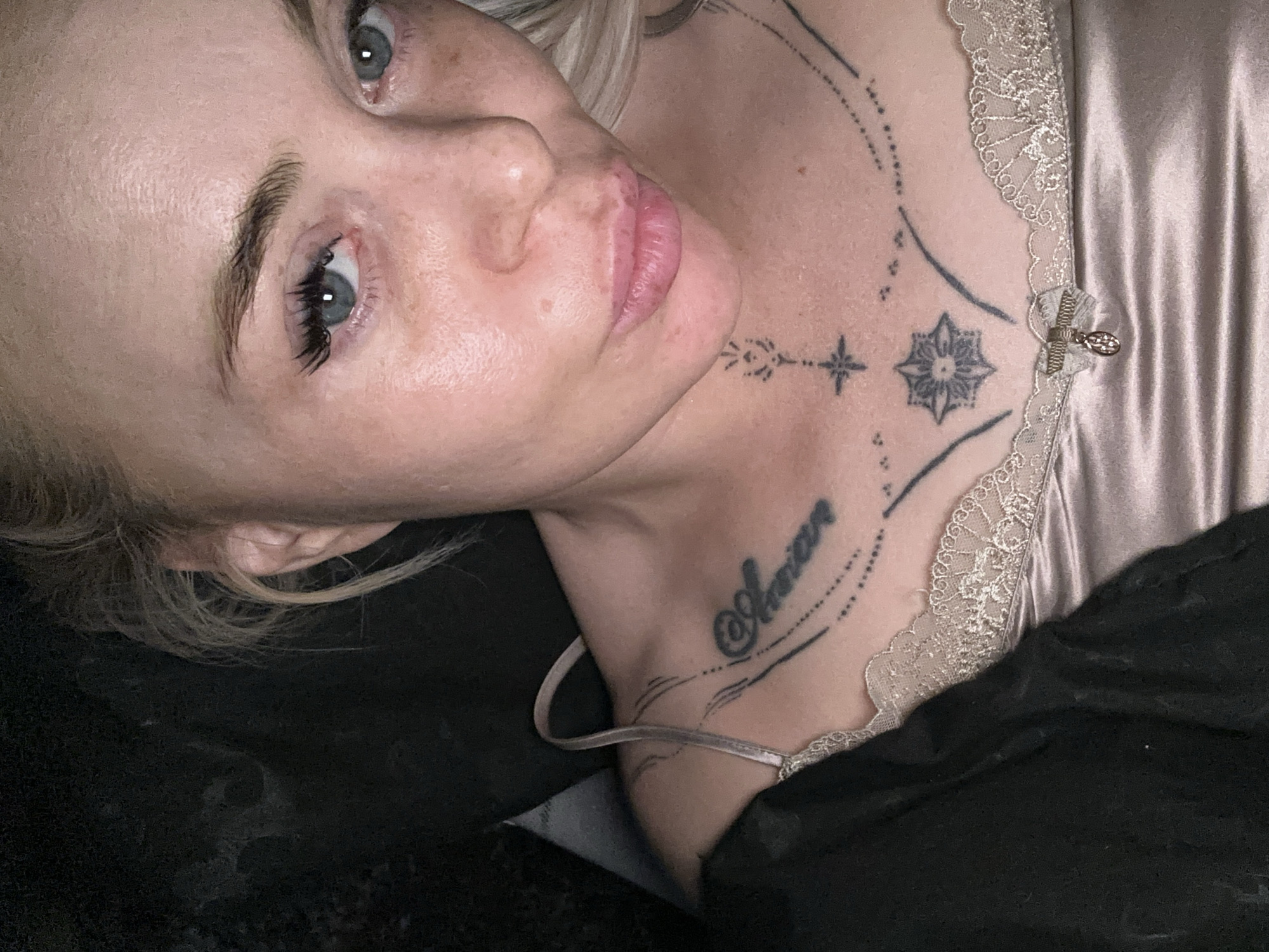 A close-up, high-angle portrait shows a blonde woman with blue eyes gazing directly at the viewer, located in Unknown, Unknown. Her skin exhibits natural texture, including visible pores and light freckles. She has full, pinkish lips formed into a slight pout. Her left eye (viewer's right) features prominent false eyelashes, and her eyebrows are well-defined. Her blonde hair appears loosely styled or pulled back, with wisps visible around her right ear (viewer's left).
She wears a light-colored, possibly champagne or pale pink, satin camisole with white lace trim along the neckline. A small, decorative bow with a silver-colored charm is attached to the center front of the camisole. A dark fabric garment (e.g., jacket, blanket) partially covers her left shoulder (viewer's right).
Extensive black ink tattoos are visible on her chest and sternum. The word "Amor" is tattooed in script on her upper left chest (viewer's right), accompanied by dotted lines. A prominent stylized star or floral geometric design is centered on her sternum, with linear and dotted patterns extending around it. A smaller, linear, arrow-like design is visible on her lower throat/upper chest. The background is dark and indistinct, suggesting an indoor setting with low ambient light or a dark surface.