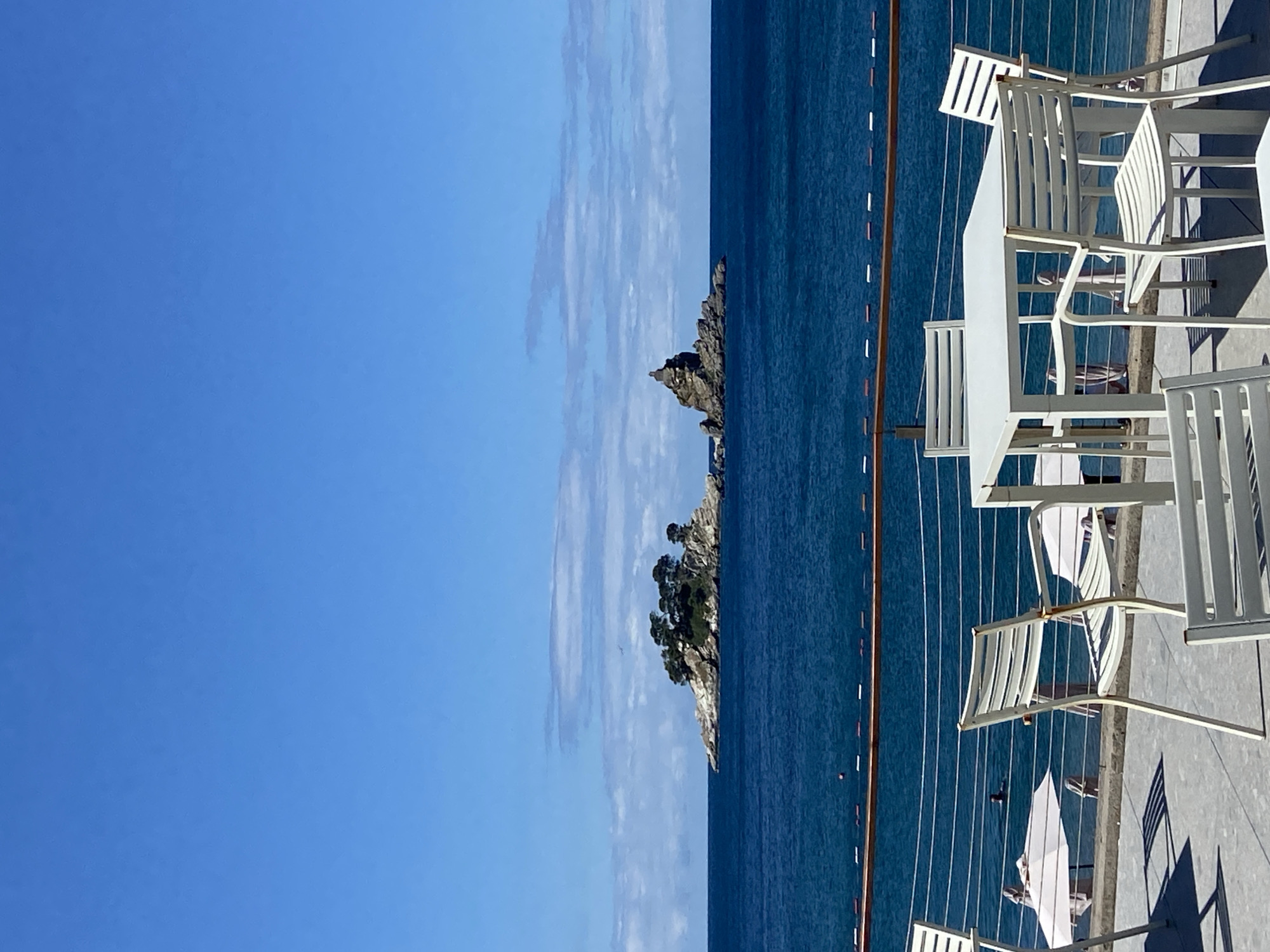 The image depicts a clear, bright day with a vast expanse of deep blue sea under a clear blue sky with scattered white clouds. In the distance, two rocky islands rise from the water. The larger island on the left is topped with lush green trees, while the island on the right is a jagged, imposing rock formation. In the foreground, a patio area is set up with white outdoor furniture, including several tables and chairs, positioned behind a railing overlooking the sea. A beach area with white umbrellas and beachgoers is visible below the patio. A person is swimming in the sea. The scene suggests a serene coastal setting.