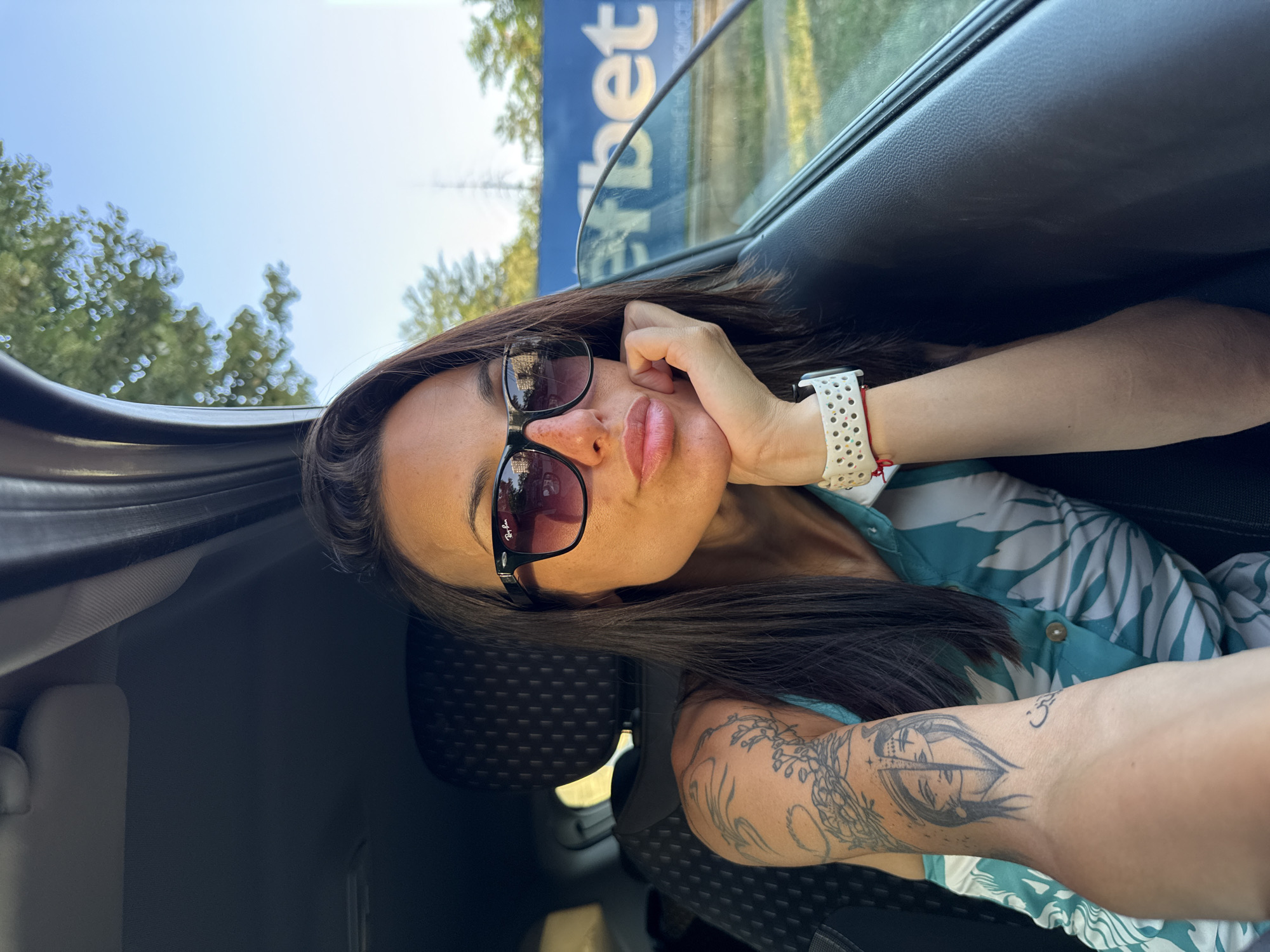 This is a selfie taken by a woman sitting in the passenger seat of a car, with the camera angled to capture her face and part of her arm and torso. She is wearing sunglasses and has her hand resting on her cheek, with her lips slightly parted. Her hair is dark and long, framing her face. She is wearing a blue and white patterned shirt and a white watch with a red string bracelet. A tattoo is visible on her arm.
The background shows trees and a blue sign with white text that is partially obscured but appears to say "eet". The photo is taken outdoors, likely during the day, with bright lighting suggesting it's daytime. The overall mood seems relaxed and casual. The location context is Pleven, Bulgaria.