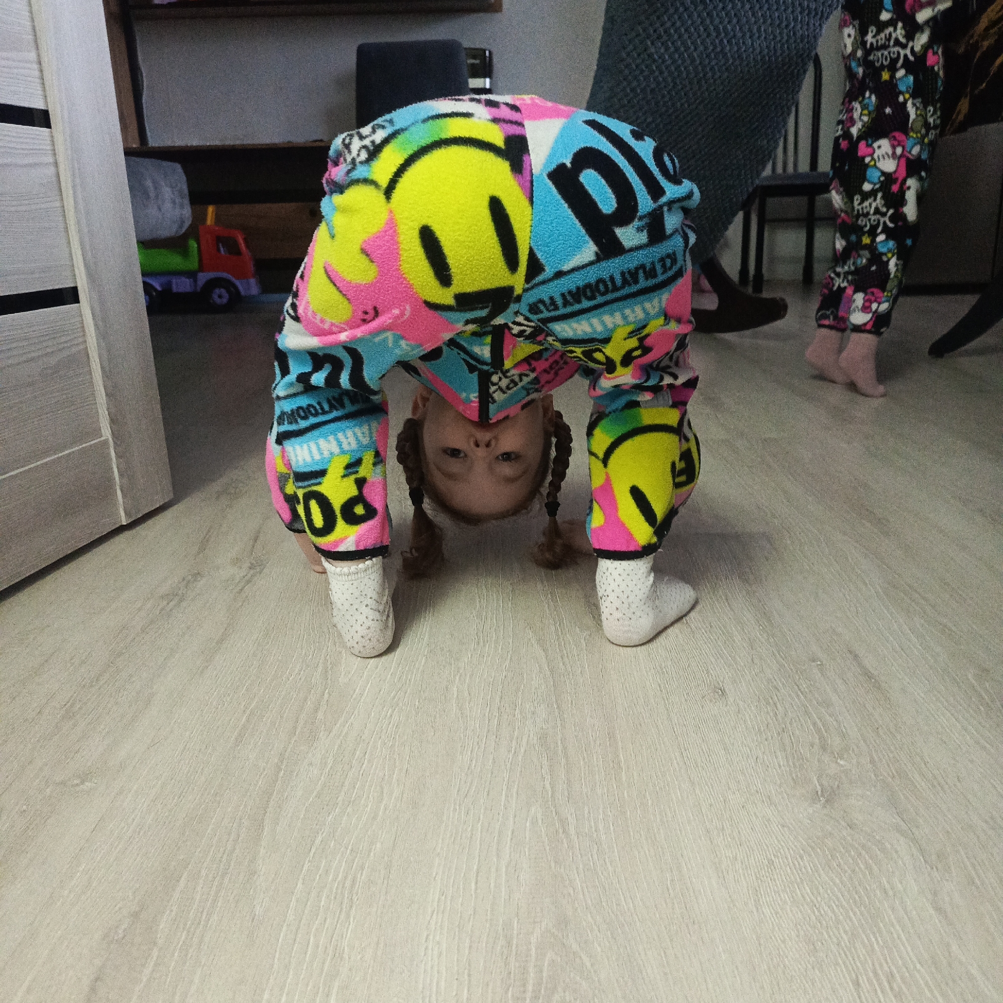 A young girl in pajamas with a smiley face and text pattern is doing a backbend on a light wood-grain floor. She is wearing white socks and has braided hair. In the background, there is a door, a toy truck, a chair, and another person wearing patterned pajamas. The location is presumably in a home or residence in Марковский, Russia.