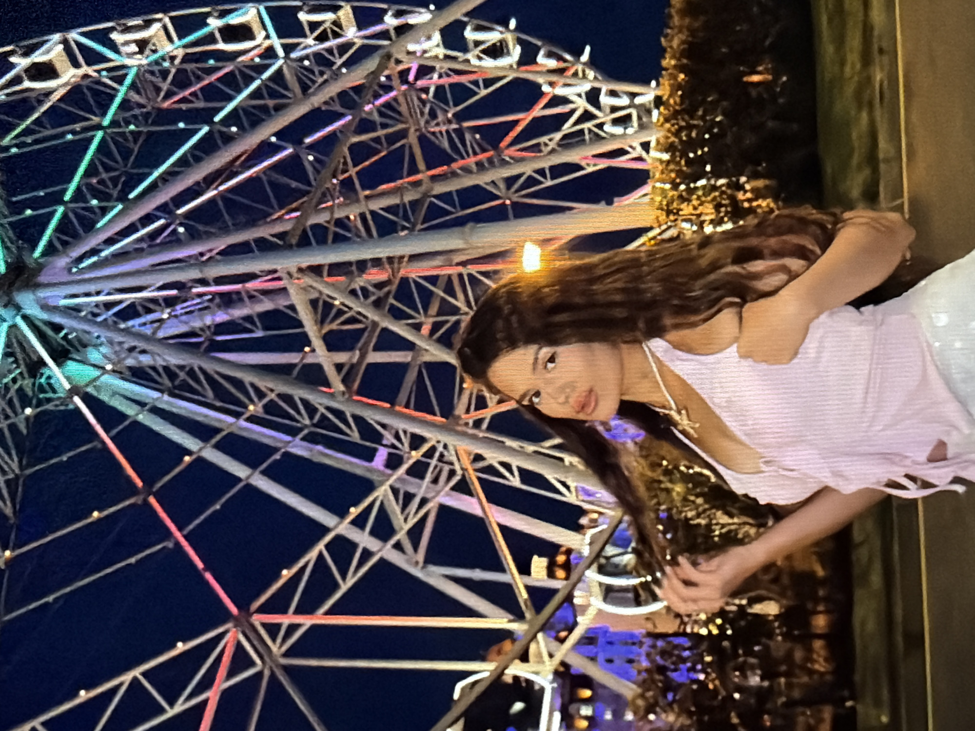 A young woman with long, dark hair is captured in a slightly tilted, medium shot, posing in front of a brightly lit Ferris wheel at night. She is wearing a light pink, textured top with ripped detailing along the hem and a delicate star pendant necklace. Her arms are crossed, and her hands are intertwined, resting near her chest. Her gaze is directed towards the viewer, with a soft, almost pensive expression.
The Ferris wheel dominates the background, its metallic structure illuminated by a vibrant array of colorful lights—greens, blues, purples, and reds—creating a dazzling spectacle against the dark night sky. Small, white lights are also visible along the spokes of the wheel. To the right of the woman, a dark, textured object, possibly a tree or a Christmas tree, is adorned with numerous small, twinkling golden lights, further enhancing the festive atmosphere. In the far distance, blurred city lights are visible, suggesting an urban or amusement park setting.
The time of day is clearly nighttime, evidenced by the dark sky and the artificial lighting. The weather appears clear, as there are no visible signs of rain or clouds. The overall mood is one of enchantment and wonder, amplified by the vibrant lights and the woman's serene presence. There is no visible text in the image.