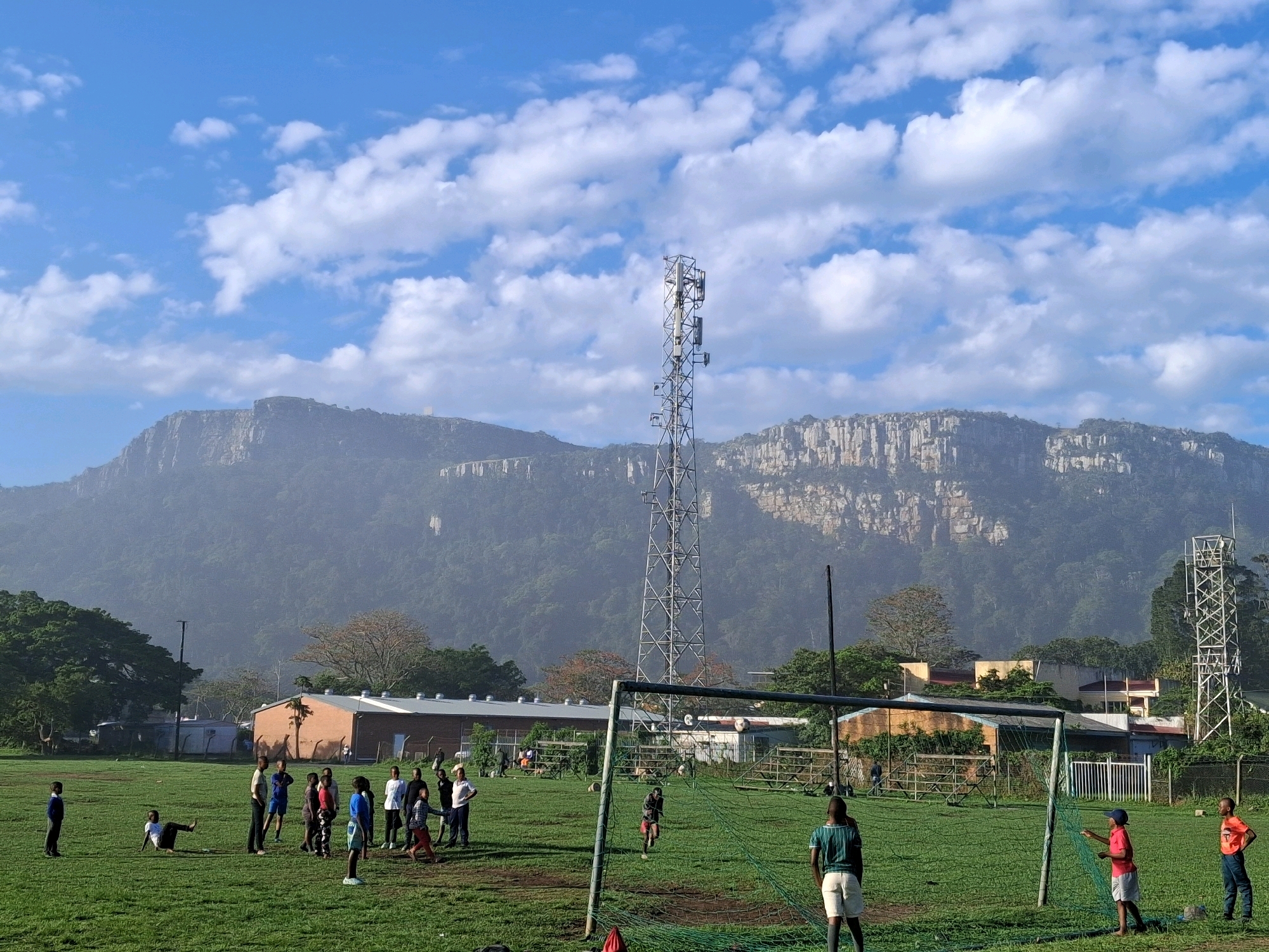 A group of children are playing soccer on a grassy field in Port St. Johns, South Africa. The field is located in front of a mountainous backdrop with rocky cliffs and lush green vegetation. A tall communication tower stands prominently in the middle ground, with various buildings and a fence visible behind the field. A soccer goal with a green net is positioned on the right side of the frame. The sky is blue with scattered white clouds.