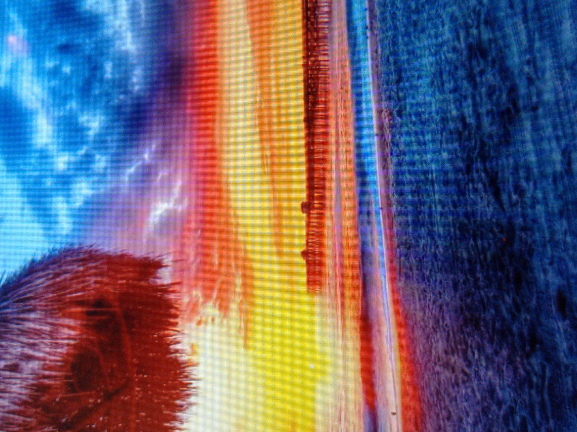 This is a low-resolution, heavily pixelated image of a sunset at a beach. A thatched umbrella or roof is visible in the upper left corner. The sky is a dramatic mix of deep blue, purple, red, and bright yellow, with visible clouds. A long pier extends into the ocean, silhouetted against the vibrant sunset. The ocean has small waves crashing on the shore. The beach in the foreground is covered in what appears to be sand, rendered in dark blue and purple hues. The image does not depict any elements that would suggest a location in Mukachevo, Ukraine, which is an inland city.