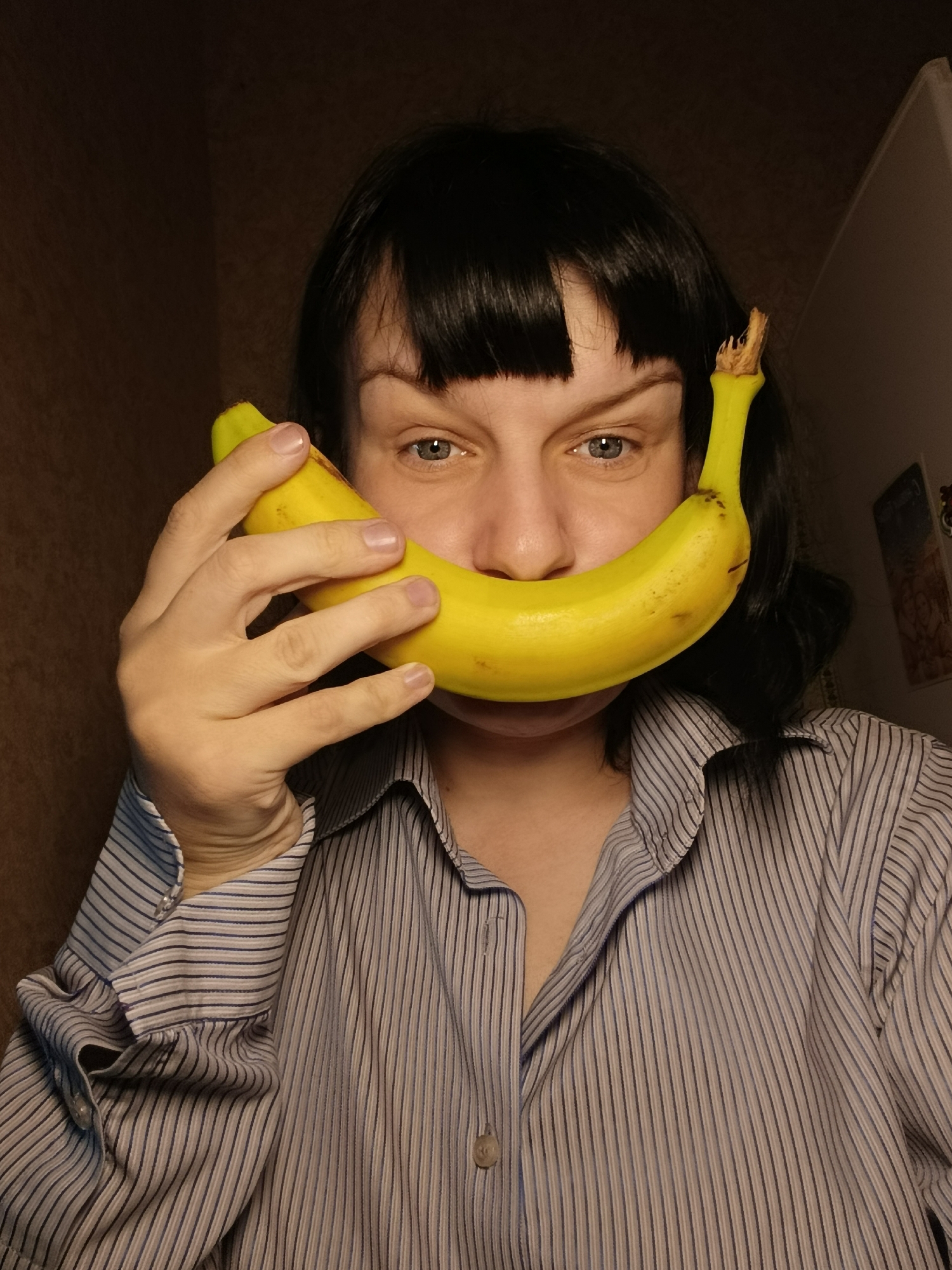 A person with dark hair is holding a banana in front of their face, mimicking a smile. The person is wearing a button-down striped shirt. The background is a dimly lit room. There is no indication of the location being Фоки, Russia.