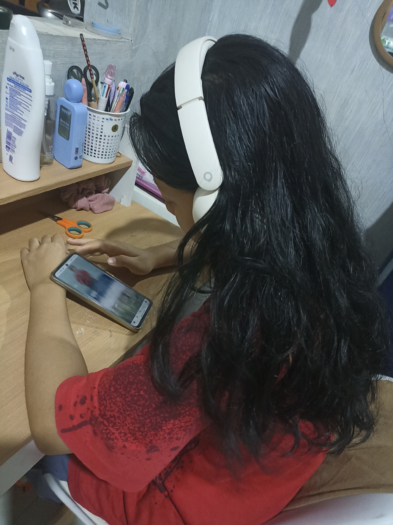 A young person, seen from behind and slightly to the side, sits at a wooden desk wearing white headphones and looking at a smartphone. The person has long, dark, wavy hair. They are wearing a red t-shirt with a distressed pattern. On the desk, in front of the person, lies the smartphone displaying a video or image of a person in a red shirt. To the left of the person, on a shelf above the desk, are various desk supplies including a white lotion bottle, a blue dispenser, a basket filled with pens and scissors, and a pair of orange-handled scissors on the desk itself. The background is a textured grey wall. The scene appears to be indoors, likely a personal workspace or study area.