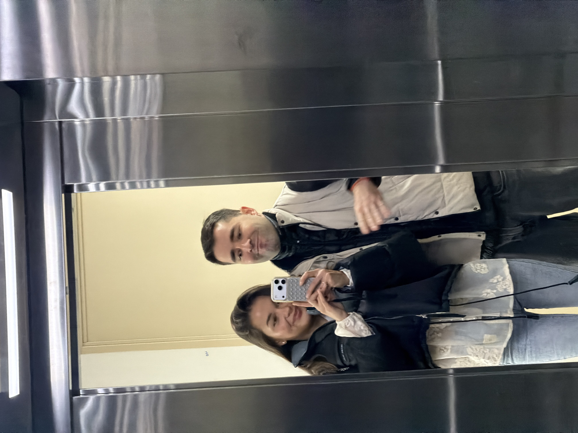 The scene depicts two individuals reflected in a polished metallic surface, likely an elevator interior or a large mirror, within an indoor environment in Tbilisi, Georgia.
On the left, a woman with long, dark hair is smiling, wearing a black puffer jacket over a light-colored, patterned blouse and light blue jeans. She holds a silver smartphone, equipped with a grey textured case, in both hands, angled towards the reflective surface, indicating she is taking a selfie. A ring is visible on her left ring finger.
To her right and slightly behind her, a man with short, dark hair is reflected. He wears a dark top under a light beige puffer vest. He has a slight smirk or pout on his face, looking towards the camera. His right hand is partially visible, raised with fingers slightly spread.
The reflected background consists of a cream-colored wall and a closed light-brown door with a dark frame. A small, dark numeral "01" is visible on the reflected wall to the left of the door frame. Above the reflected wall and door, a horizontal white light fixture is present. The metallic surface itself shows characteristic wavy distortions in the reflection.