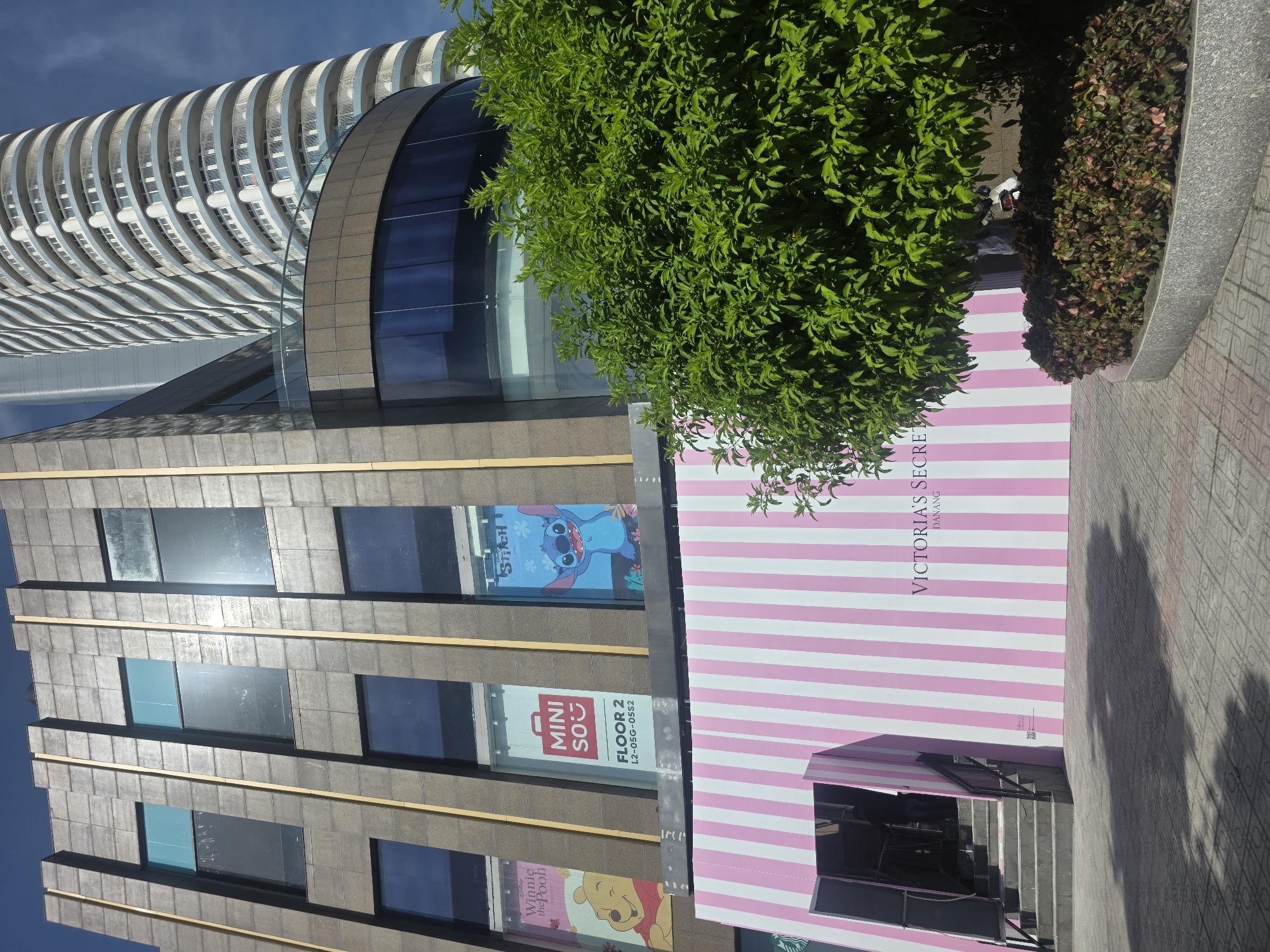 The image shows a mixed-use building with commercial and residential components in Đà Nẵng, Vietnam. The commercial section of the building is visible with several storefront signs. One of the storefronts displays a pink and white striped facade, with the words "VICTORIA'S SECRET DANANG" visible vertically. Adjacent to it, other storefronts advertise "MINI SO" on Floor 2, and cartoon character branding. The residential part of the building is a modern high-rise with curved balconies. The environment appears to be an urban area with sidewalks and landscaping. The image's orientation is turned on its side.