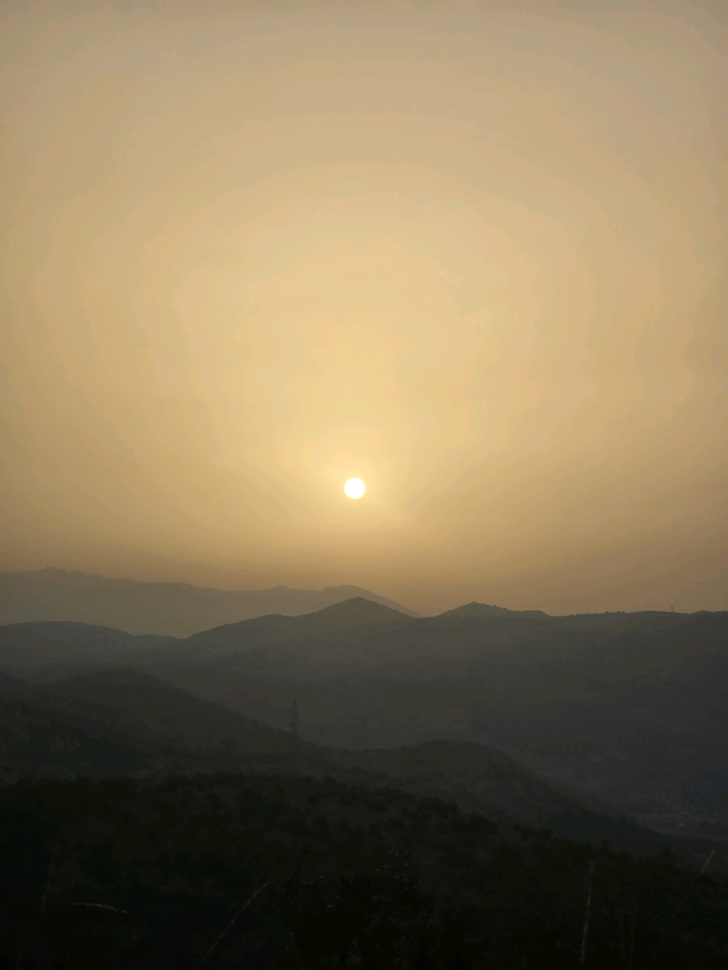 A hazy, pale yellow sky dominates the upper two-thirds of the frame, with the sun, a bright white circle, positioned slightly left of center. Below the sky, a series of silhouetted mountain ranges recede into the distance, their contours softened by atmospheric haze. The foremost mountain ridge in the foreground is dark and indistinct, with hints of vegetation. A tall, thin electrical pylon is visible on one of the mid-ground ridges. The overall impression is of a sunrise or sunset over a mountainous landscape, possibly near Podgorica, Montenegro.