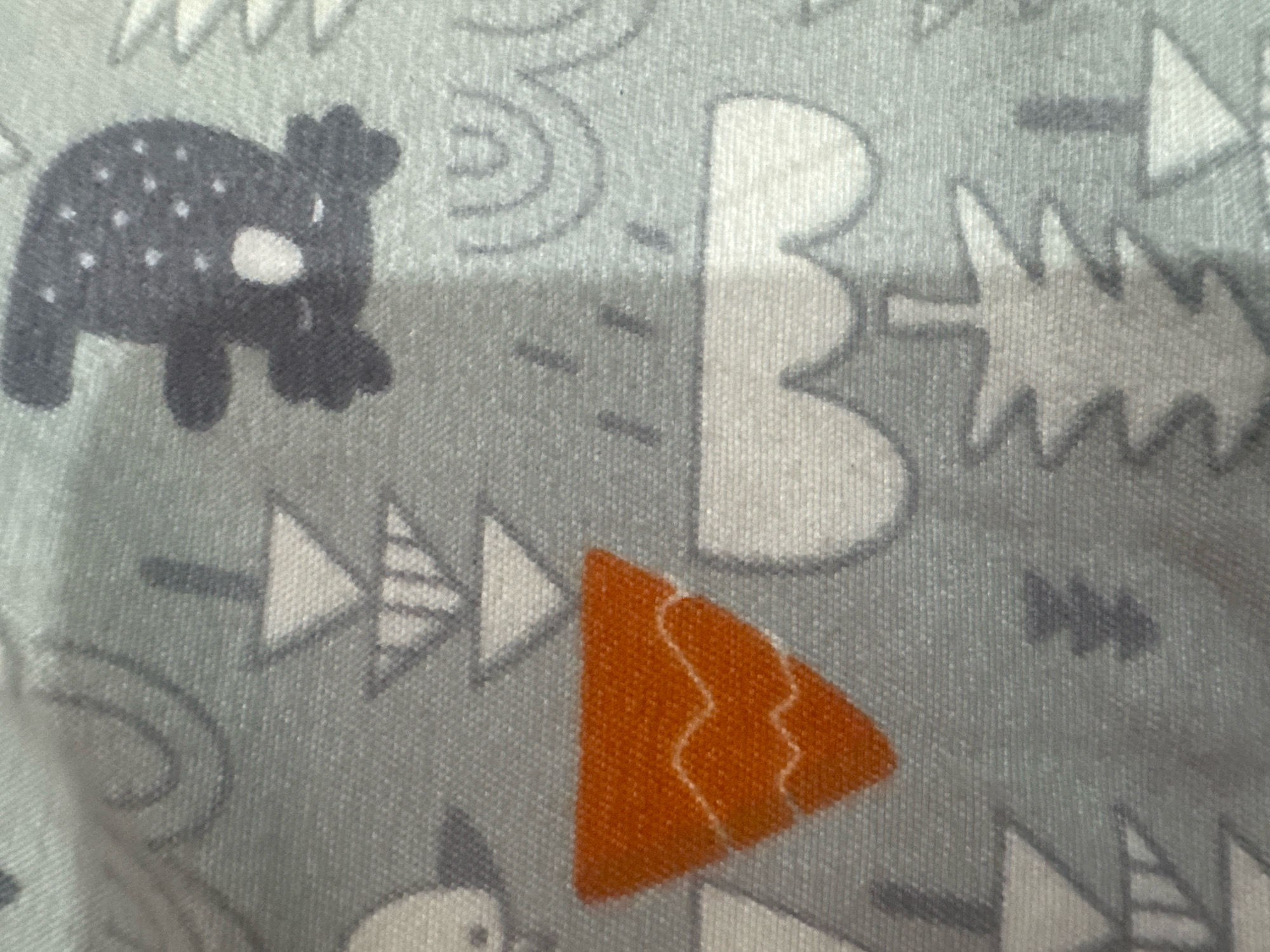 The image is a close-up of a light blue fabric with a repeating pattern of various graphic elements. The elements include a dark gray stylized bear with white polka dots and a white oval on its face, several white abstract shapes resembling clouds or stylized trees, and an orange triangular shape with white wavy lines inside. There are also several gray arrows pointing in different directions. The fabric appears to be textured, suggesting it might be cotton or a similar material, possibly from clothing or home decor. There is no identifiable location in the city of Rufisque, Senegal, as the image is a fabric pattern.