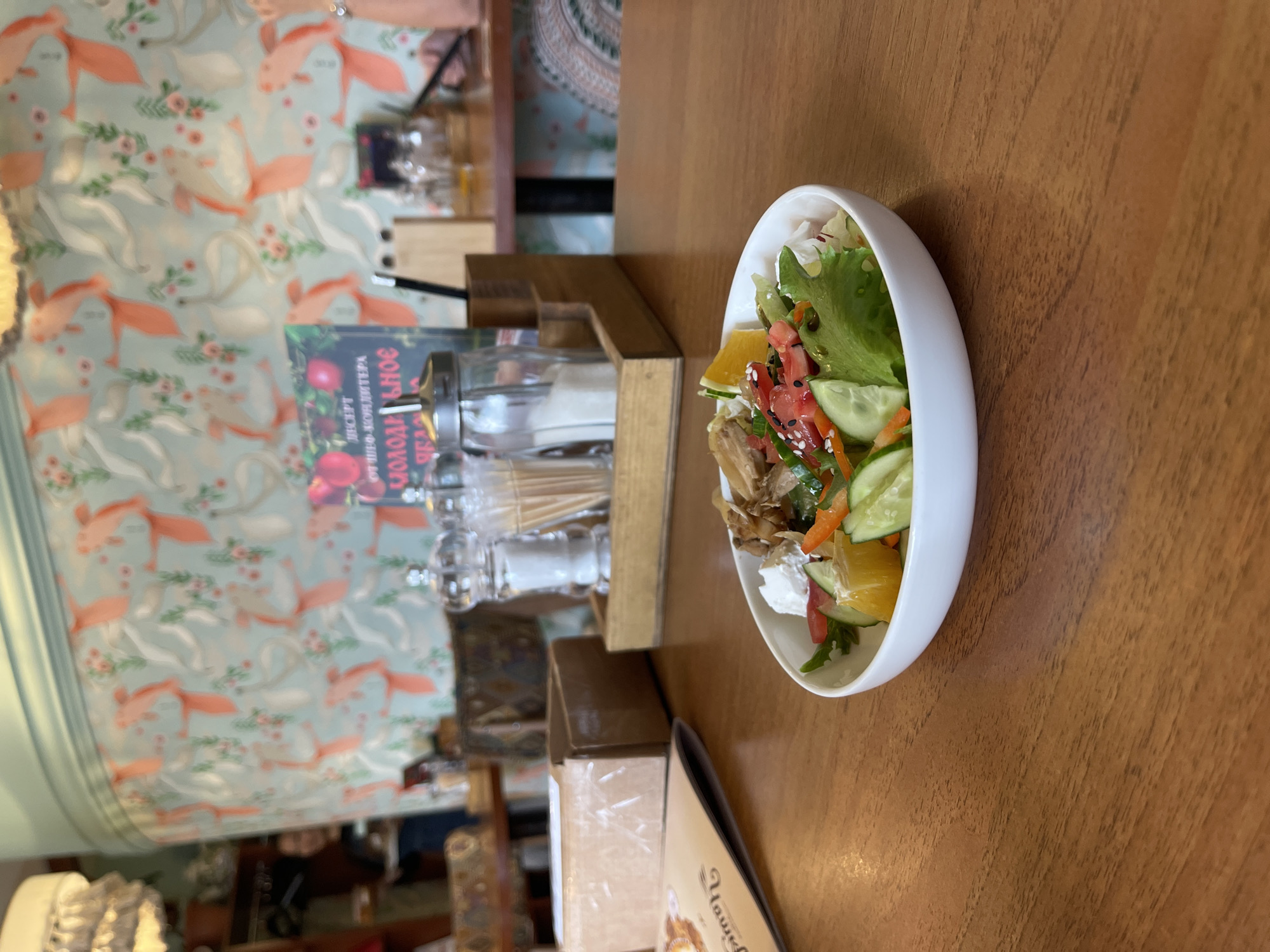A top-down, slightly angled medium shot captures a salad in a white bowl, positioned on a wooden table. The salad appears to be a mix of greens, cucumber slices, orange segments, tomatoes, and what looks like shredded chicken or fish, possibly topped with feta cheese and sesame seeds. To the left of the salad, a wooden condiment holder contains salt and pepper shakers and toothpicks. Behind the condiment holder, a book with red and green elements and Cyrillic text is visible. The background features a wall with a light blue wallpaper adorned with a pattern of stylized pink birds and green foliage. The overall setting suggests a casual dining environment, possibly a cafe or restaurant. The lighting seems to be indoor, ambient light, with no strong shadows indicating a specific time of day. There are no visible people in the frame.
Visible text on the book appears to be in Cyrillic and reads "СЛАДКОЕ и СОЛЕНОЕ" on the spine and potentially "ИНСПЕКТ КОНДИТЕРА" or similar on the cover, suggesting it might be a cookbook. The overall mood is calm and inviting.