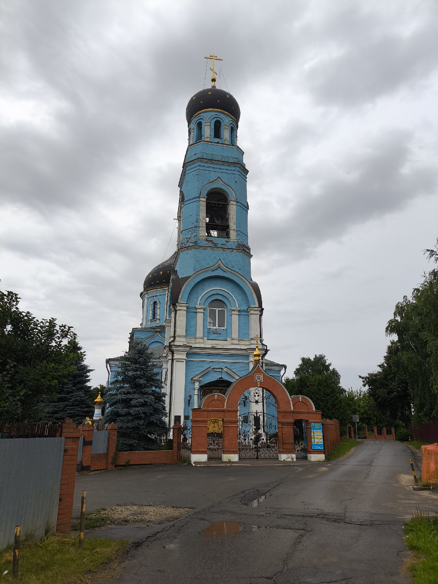 An exterior, eye-level, medium shot captures a bright blue Orthodox church under a heavily clouded sky. The church features a prominent bell tower topped with a dark dome and a golden cross. A smaller, dark dome with golden stars sits atop the main church structure. Red brick walls surround the church, with an arched gateway featuring black metal gates. Trees, including evergreen and deciduous varieties, flank the church and its grounds. The foreground shows a cracked asphalt road with puddles, and a metal fence to the left. The church is identified as the Church of the Intercession of the Most Holy Theotokos in the city of Pokrov, Russia.