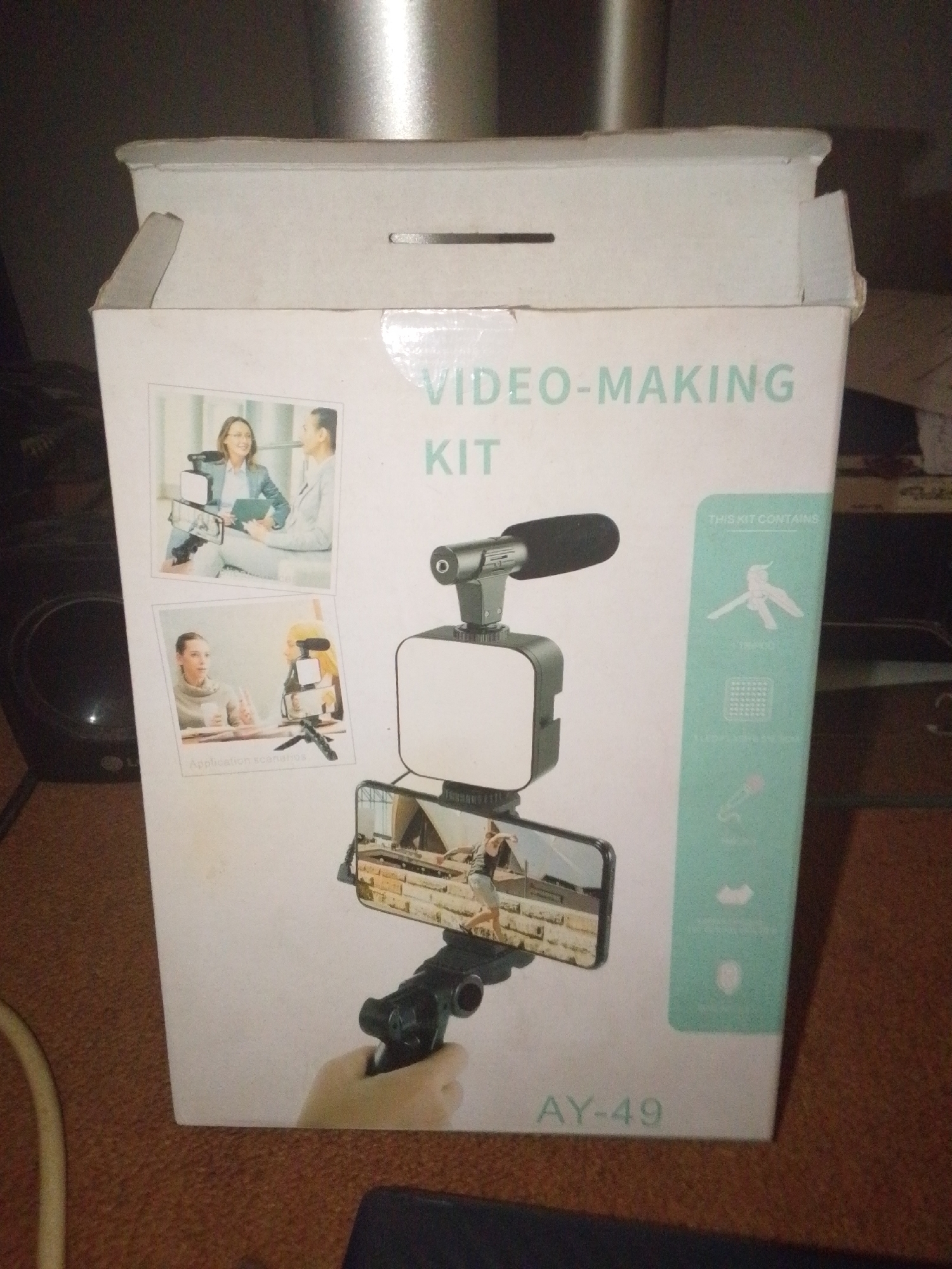 The image shows the front of a white cardboard box for a "VIDEO-MAKING KIT" labeled "AY-49". The box is slightly damaged at the top.
The front of the box features several images. The largest image shows a smartphone mounted on a hand-held gimbal with a small LED light and a microphone attached. The phone screen displays a video recording of a person walking outdoors.
Above the main image, two smaller square images depict application scenarios. The top-left image shows a woman and a man sitting and talking, with a camera setup between them. The bottom-left image shows two women talking, with a similar camera setup on a table.
To the right of the main image, a light green section lists the contents of the kit with icons and text. Visible text includes "THIS KIT CONTAINS", "TRIPOD", "LEDGE LIGHTING USE", and an icon of a microphone.
The background of the image is a dark, textured surface, possibly a carpet, with some visible electrical cords. The overall lighting is dim, suggesting an indoor setting. There are no explicit cues about the time of day, weather, or specific emotions. The location context provided is Jalingo, Nigeria, but this cannot be visually confirmed from the image itself.