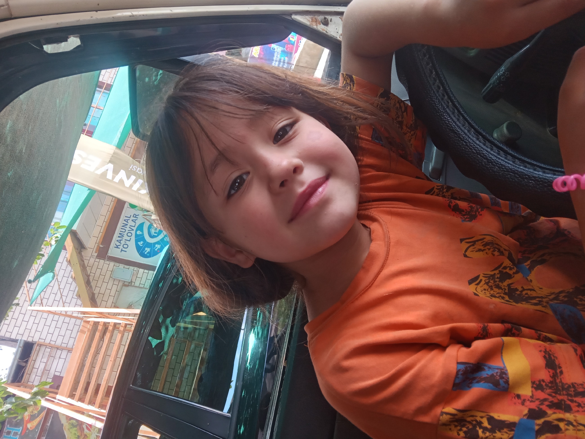 A young girl with brown hair and dark eyes smiles at the camera, leaning against a steering wheel inside a vehicle. She is wearing an orange t-shirt with a colorful, abstract pattern. Her left arm is bent, resting on the steering wheel, and her right arm is visible on the right side of the frame. The interior of the vehicle appears to be old or worn, with a grey dashboard. Outside the car, through the windows, a building with a brick facade and light-colored horizontal siding is visible. A banner with text, partially obscured and rotated, hangs on the building. Green foliage is also visible. The lighting suggests it is daytime. The text on the banner appears to be in a non-Latin script and includes the word "INVEST". Another sign with blue and white text and icons is visible. The scene appears to be in Obizarang, Uzbekistan, based on the location context. The girl's expression is happy and playful, suggesting a casual or fun moment captured inside the car.