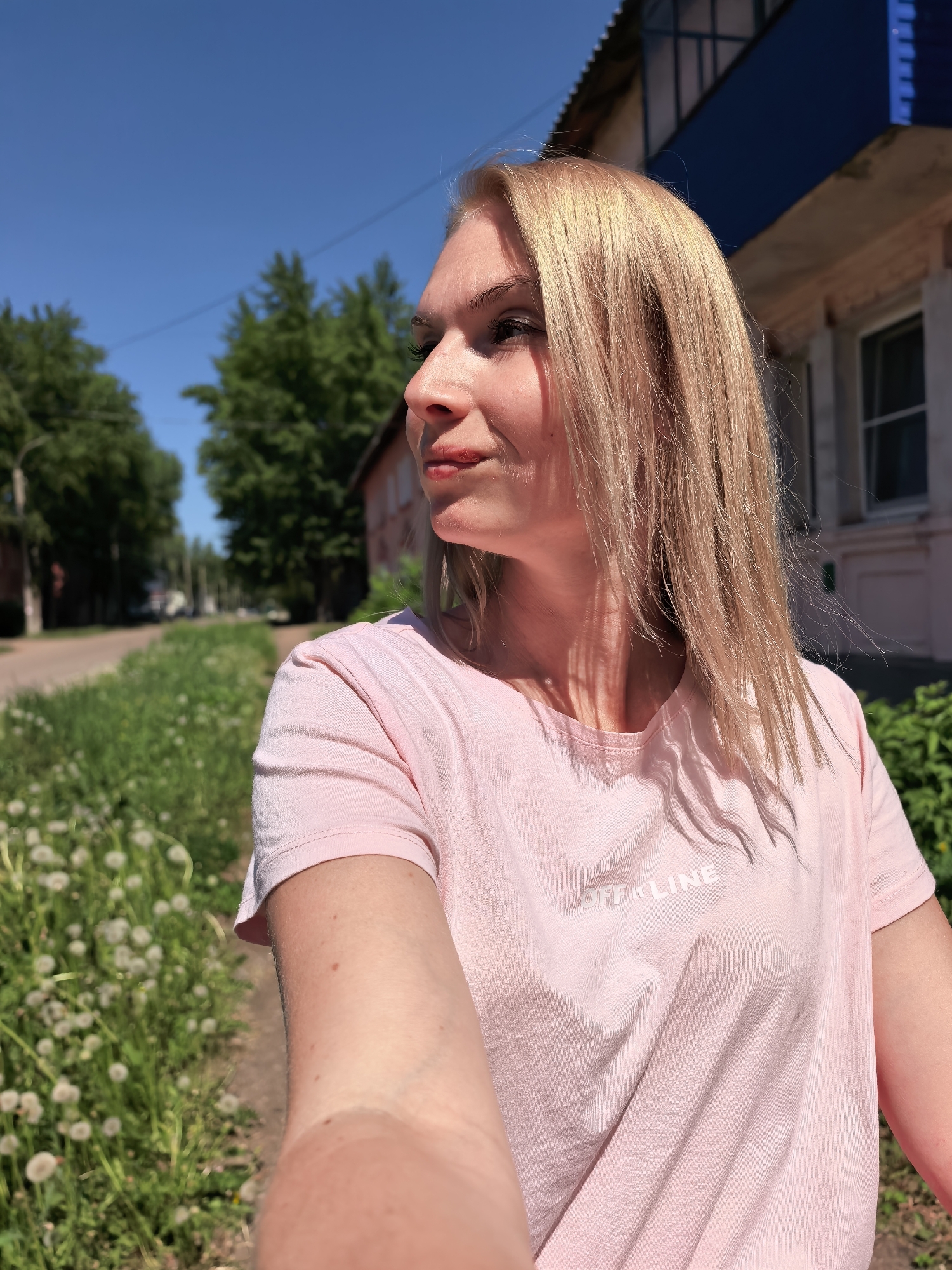 A blonde woman in a pink T-shirt stands on the side of a road with a grassy verge and dandelions. She is looking to her left. The background includes a street with trees and buildings in Sterlitamak, Russia.