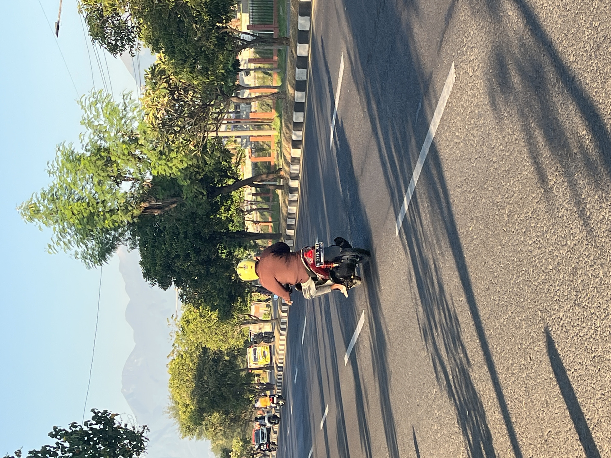 A person wearing a brown shirt and light-colored pants rides a dark-colored scooter with a red taillight down a paved road in Siring, Indonesia. The rider wears a yellow helmet. The scooter has a license plate that reads "B 2310 UZ". In the background, there are trees with green foliage, a fence, and distant mountains under a clear blue sky. Several other vehicles, including cars and scooters, are visible further down the road. The sun casts long shadows across the road.