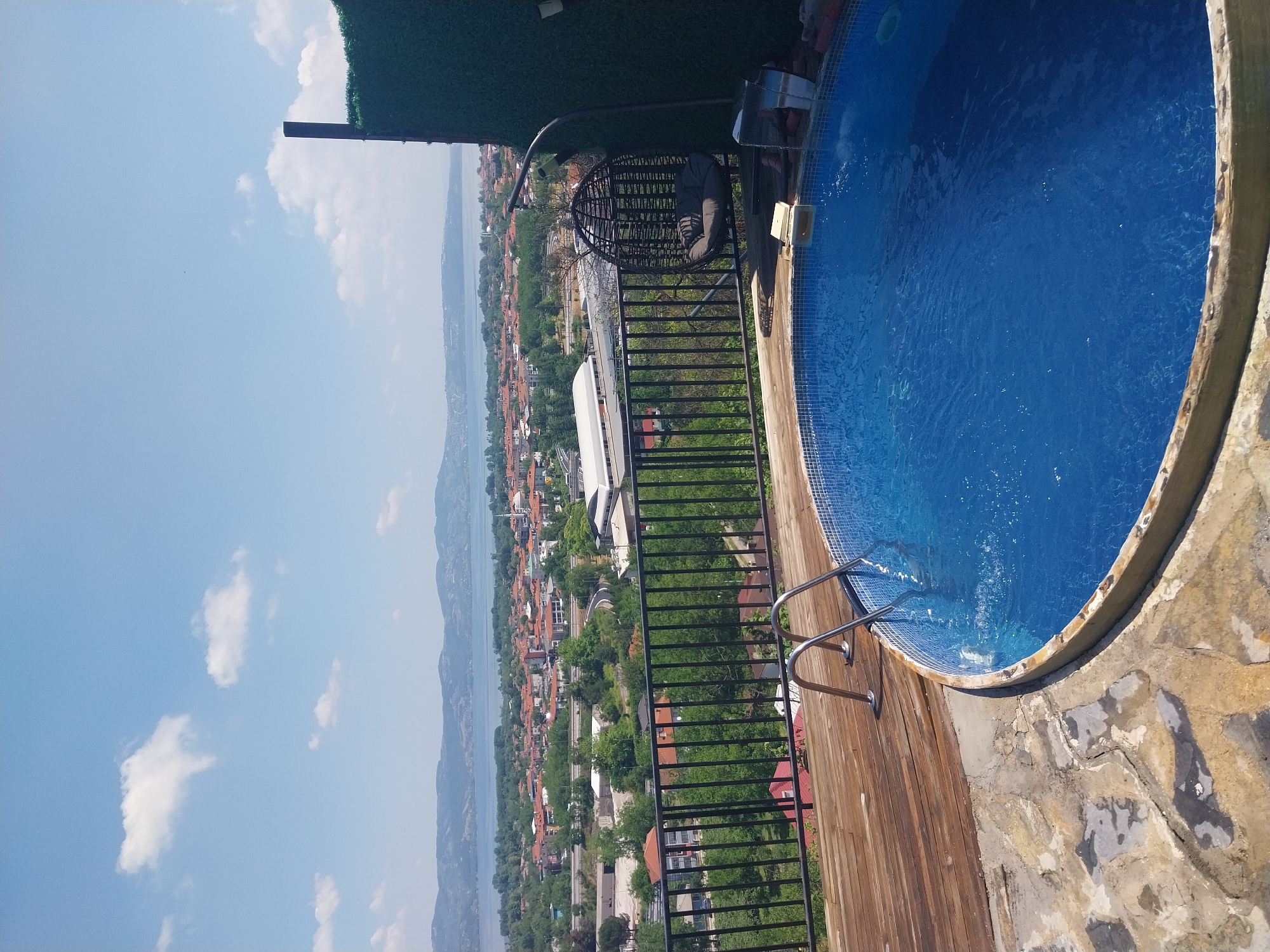 The image shows an outdoor swimming pool with clear blue water, surrounded by a wooden deck and a stone patio. A metal ladder leads into the pool. In the background, a town with red-tiled roofs is visible, situated by a body of water and rolling hills. The sky is blue with scattered white clouds. A hanging chair is positioned on the deck near a green privacy fence. The location is identified as Sapanca, Turkey. There are no people visible in the image.