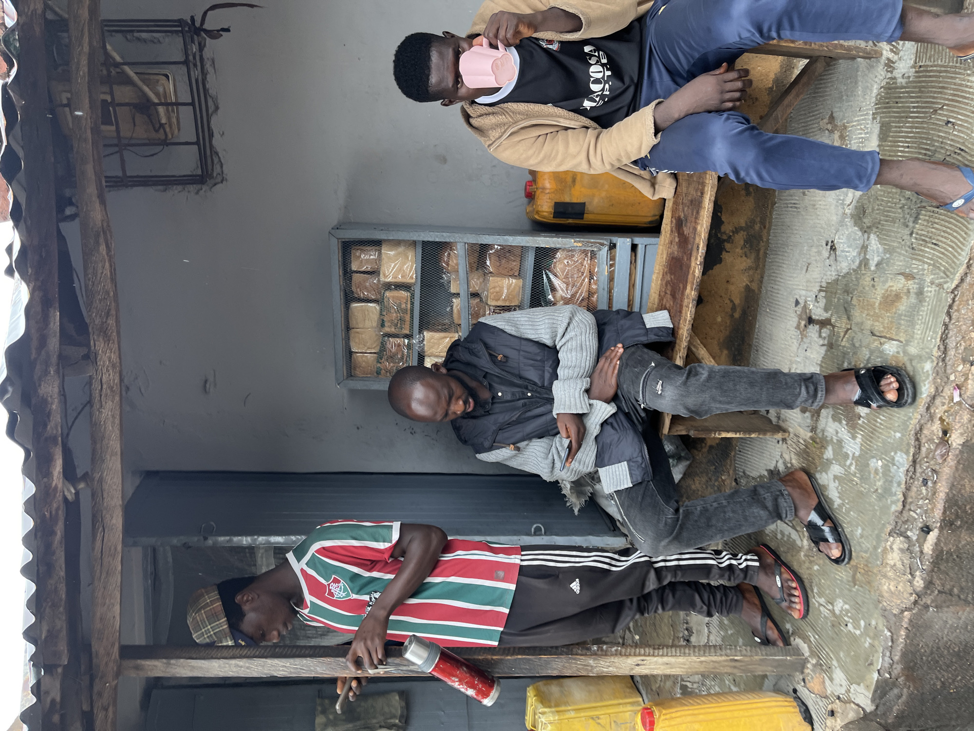 Three men are sitting on a wooden bench in front of what appears to be a bakery in Jos, Nigeria. The bakery has a display case with loaves of bread. One man is drinking from a pink mug while wearing a beige jacket and dark pants. Another man is clutching his stomach, wearing a grey sweater and black pants. The third man is working on something while holding a red thermos, wearing a red and green striped shirt and dark pants. Two yellow gas containers are visible near the ground.