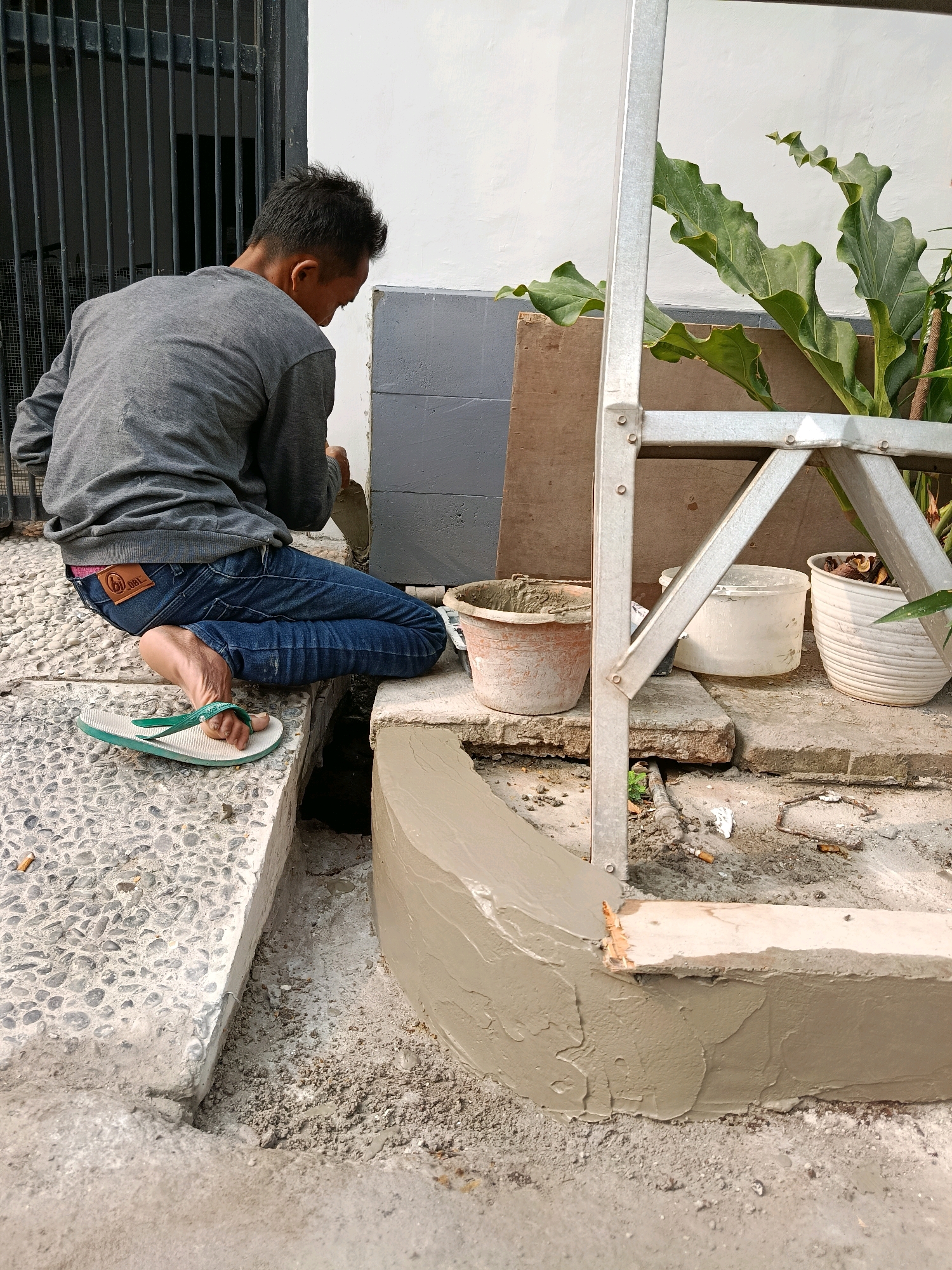 A man is kneeling on a gravel and concrete surface, wearing a grey long-sleeved shirt, blue jeans, and green flip-flops. He is applying cement to a curved concrete structure with a trowel. In the background, there is a white wall, a metal grate, a large leafy plant, and several pots, one containing cement. The scene appears to be outdoors in a residential or construction area, possibly in Bekasi, Indonesia, given the presence of what seems to be an Indonesian brand on the man's jeans.