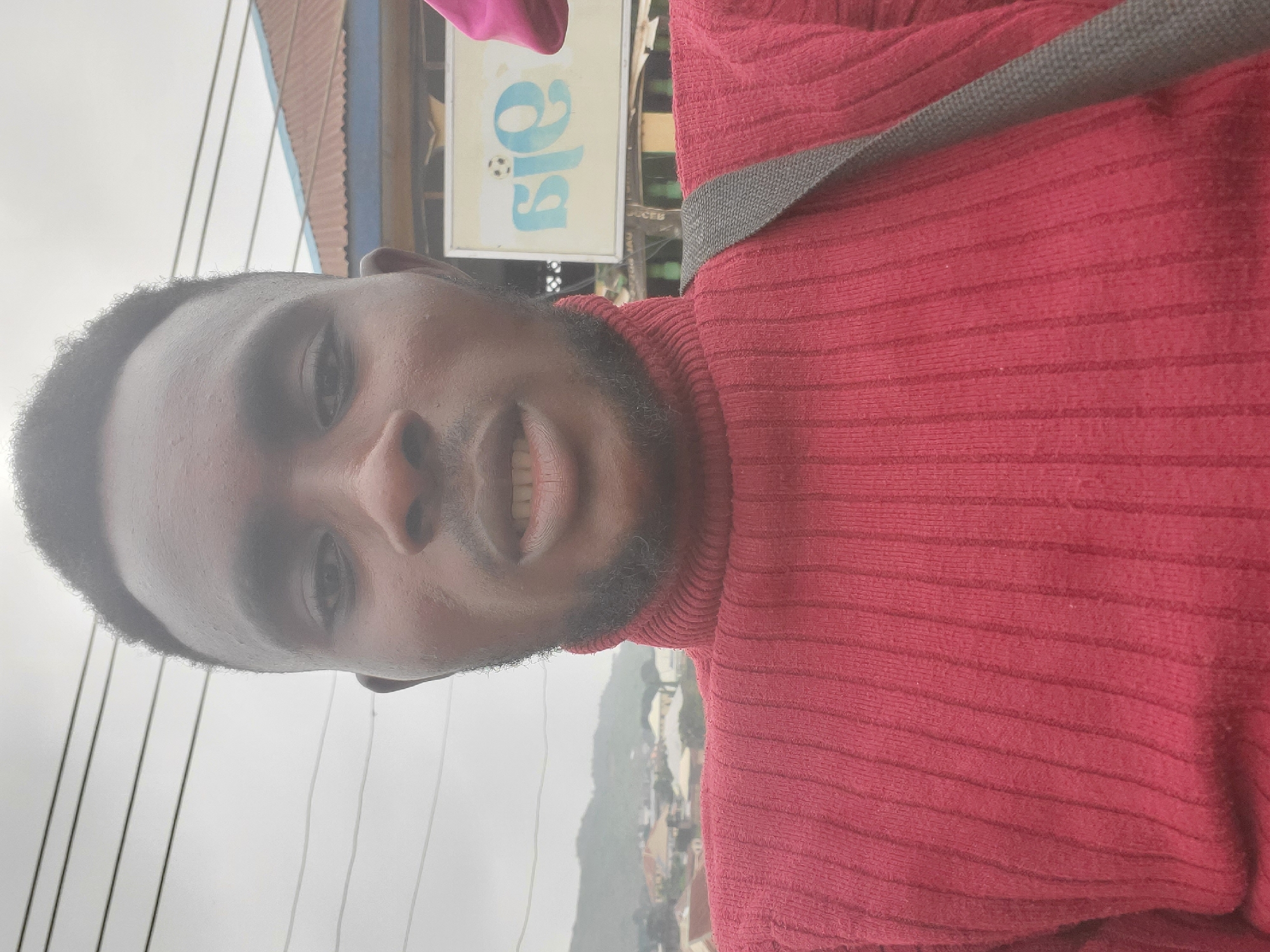 A young Black man with a short haircut and a beard is taking a selfie outdoors. He is wearing a red, ribbed turtleneck sweater and a gray strap across his shoulder. The background shows a cloudy sky with power lines, a building with a corrugated metal roof, and a sign with blue lettering and a soccer ball. In the distance, there are hills and houses, suggesting a semi-urban or town setting. The man's facial expression is neutral, and he is looking directly at the camera.