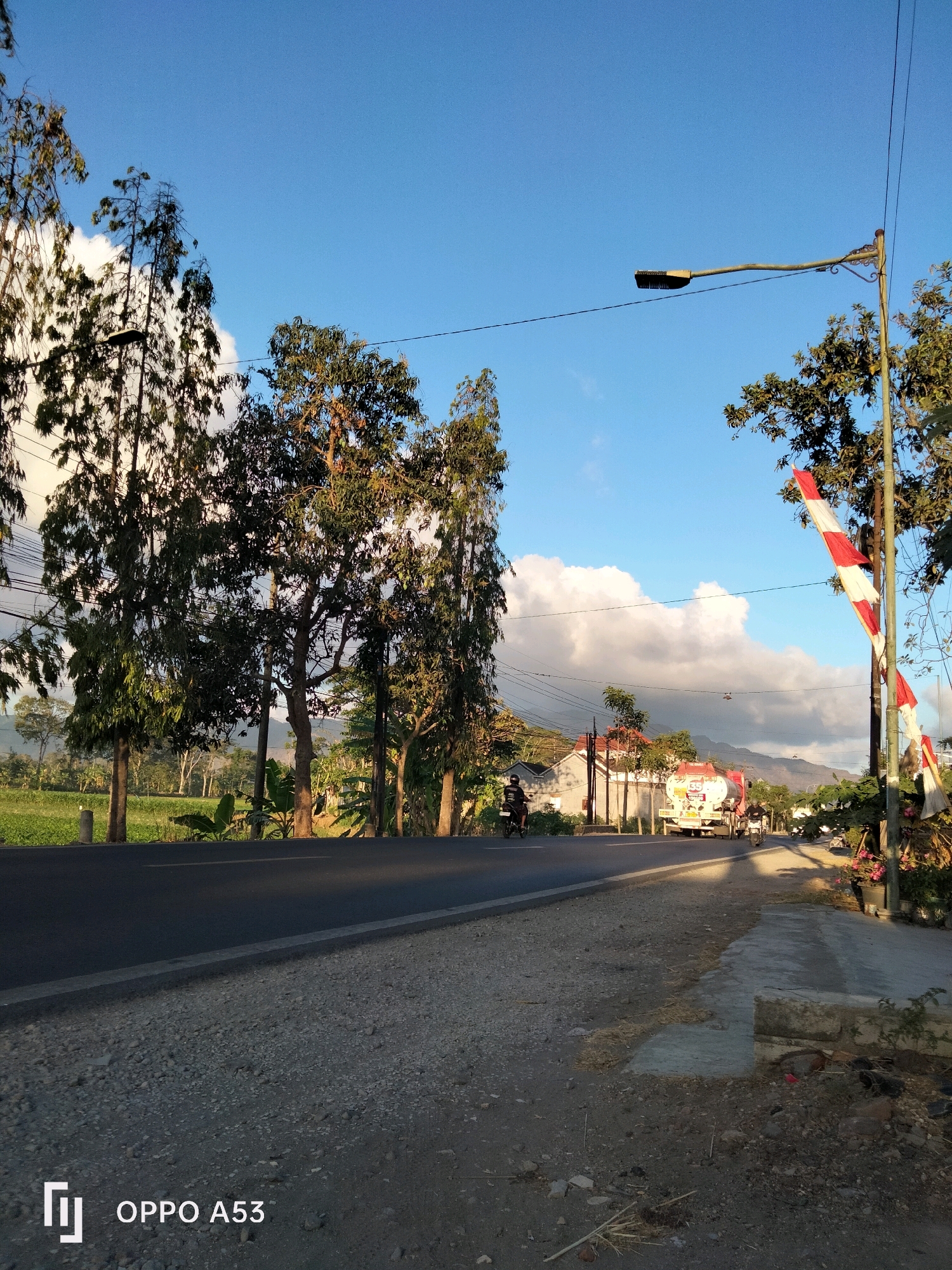 The image depicts a road scene in the city of Trenggalek, Indonesia, bathed in the golden light of a late afternoon sun. Tall trees with lush green foliage line the left side of the asphalt road, casting long shadows. On the right side, a red and white striped barrier, commonly seen at level crossings or restricted areas, is erected next to a utility pole. Further down the road, a white tanker truck with red accents is visible, approaching from the distance. Two individuals on motorcycles are also present, one in the foreground and another further ahead, navigating the roadway. The background reveals residential structures, lush green fields, and a range of hills or mountains under a bright blue sky dotted with fluffy white clouds. The foreground shows a gravelly roadside with scattered debris. The timestamp "OPPO A53" is overlaid in the bottom left corner of the image.