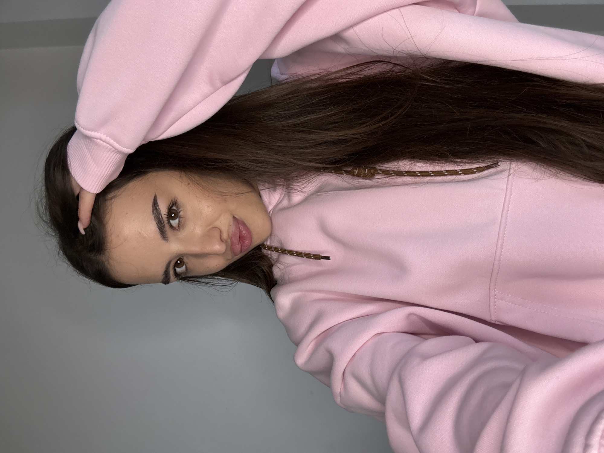 The image is a close-up selfie of a young woman with long, dark brown hair, wearing a pink hooded sweatshirt. She is indoors, against a plain gray wall. Her right hand is resting on her head, and she is looking towards the camera with her lips slightly pursed. The lighting is soft and even, suggesting an indoor setting with artificial light. There are no discernible landmarks or features that would allow for the identification of a specific location within Chișinău, Moldova.