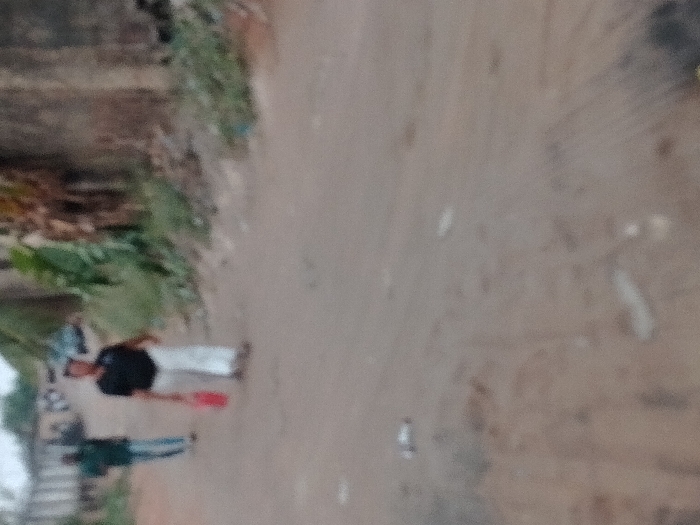 The scene depicts an unpaved dirt road in Orie Uratta, Nigeria, under an overcast sky. In the mid-ground, a male individual, dressed in a dark shirt and light-colored trousers, walks towards the left, holding a red object in his right hand. Further down the road, another individual, wearing a dark top and light blue trousers, walks in the same general direction. The right side of the road is bordered by green foliage, some dried brown plant matter, and a textured concrete or mud wall. The dirt road surface is light brown, uneven, and features scattered white and light-colored debris. In the far background, multiple vehicles, including a large truck or bus, and indistinct structures are visible. No direct interaction between the individuals is observed.