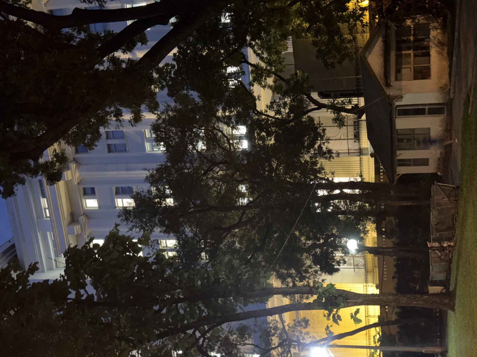 This is a nighttime photograph of a building in Hanoi, Vietnam, viewed from across a grassy area with trees in the foreground. The building appears to be a large, multi-story structure with white columns and numerous lit windows. To the right of the building, there is a small, white, single-story structure with a dark roof, possibly a guardhouse. A blurred figure is walking past this structure. In the middle ground, there are several metal bins or carts on wheels. The scene is illuminated by streetlights and the lights from within the building.