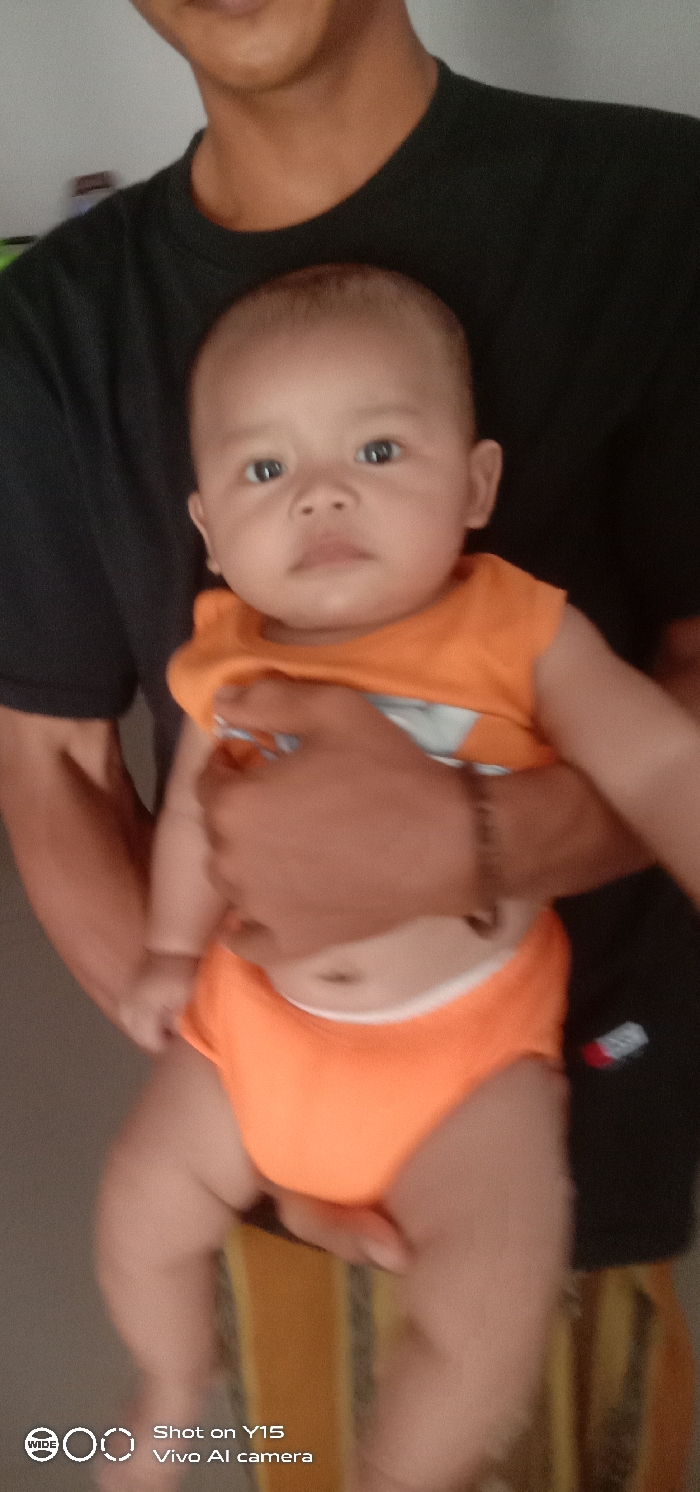 An adult, mostly cropped, holds an infant in an indoor setting within Brumbungan Kidul, Indonesia. The adult's visible features include a medium-toned lower face (mouth and chin), neck, and the right arm, which supports the infant's lower torso. The adult wears a black short-sleeved t-shirt and a dark, possibly braided, bracelet on the right wrist. The infant, centrally positioned and facing the camera, has light brown skin, dark eyes, and a neutral expression. The infant is dressed in an orange sleeveless top with a light-colored graphic design on the chest and orange bottoms. The background consists primarily of a light-colored, plain surface, with a vertical object featuring yellow and dark stripes partially visible in the lower right. A digital watermark in the lower left indicates "Shot on Y15 Vivo AI camera."