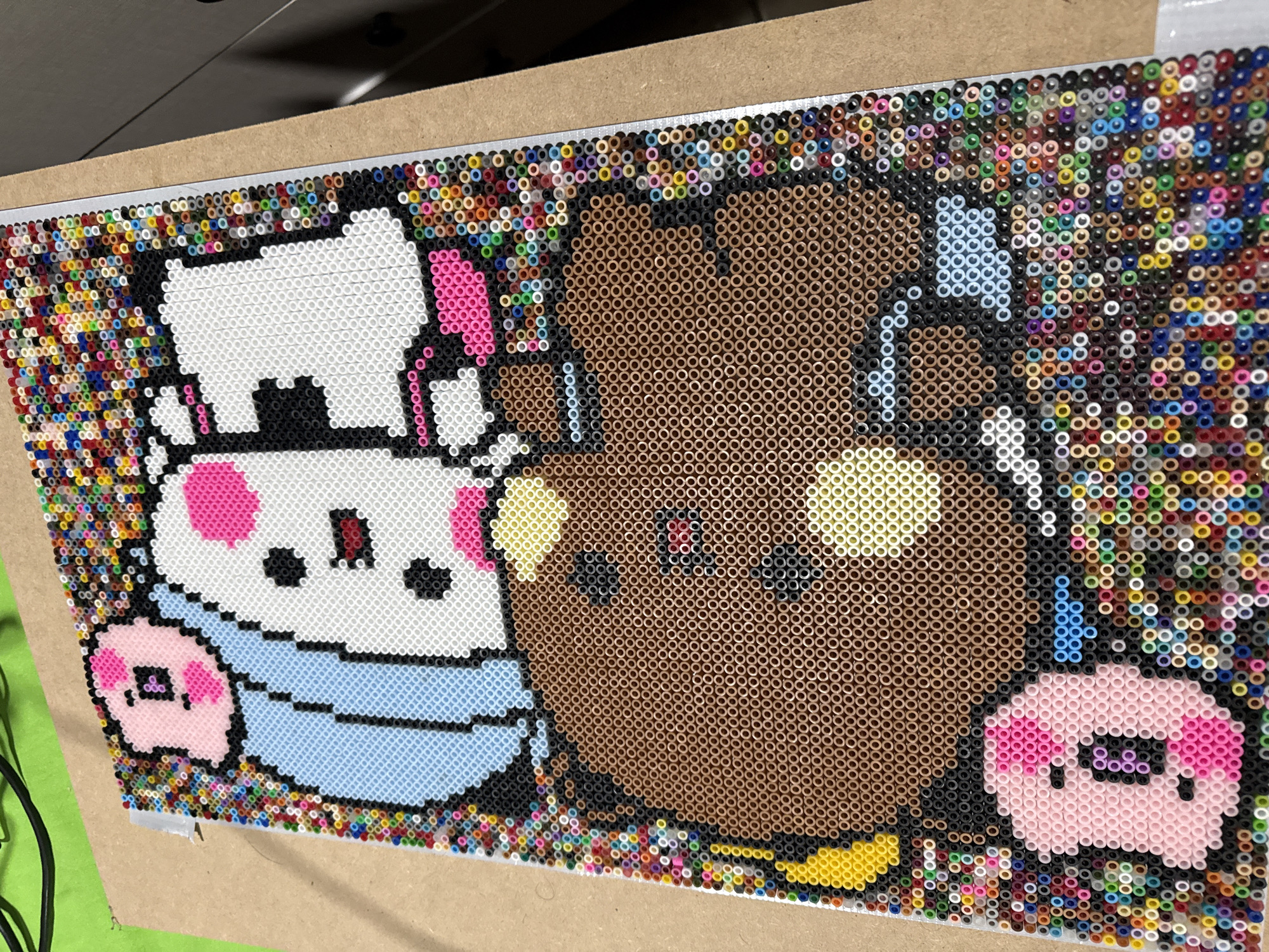 This image shows a craft project made from Perler beads depicting cartoon characters. On the left, there is a white character wearing a light blue bib-like garment, with a pink detail and a small pink character beside it. To the right, there is a brown bear character with yellow details and a pink character to the right of it. The Perler bead design is set on a board. The background is filled with a colorful pattern of Perler beads. The location is unknown as there are no identifying details such as street signs, landmarks, or distinctive architectural styles visible. The city of Puebla, Mexico is not supported by the information in the image.